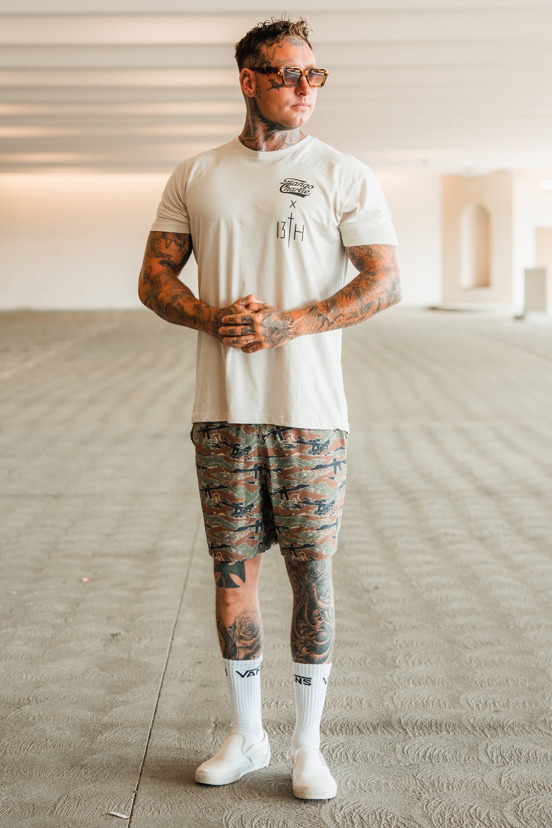 A tattooed man in sunglasses and camouflage shorts wears the Tango Charlie Apparel Lift Heavy. Pray Hard. tee, standing indoors on a textured floor with hands clasped, channeling spiritual resilience as he looks to the side.