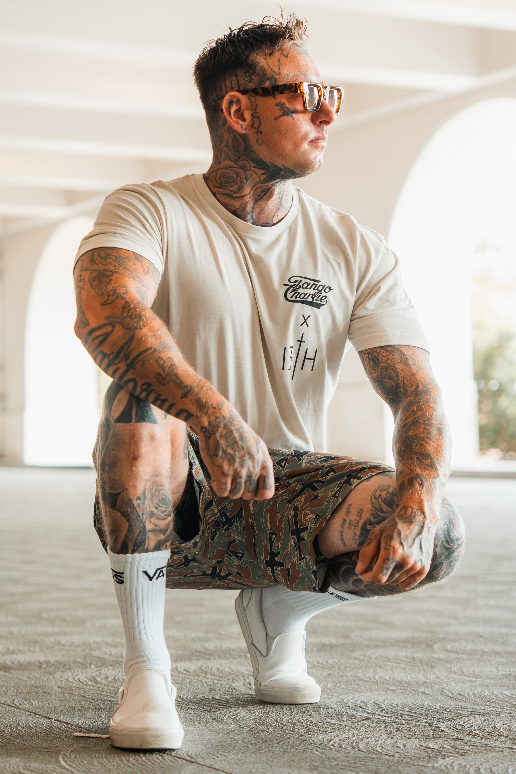 A tattooed person in Tango Charlie Apparels Lift Heavy. Pray Hard. tee, sunglasses, patterned shorts, white socks, and shoes crouches outdoors by sunlit archesâembodying spiritual resilience.