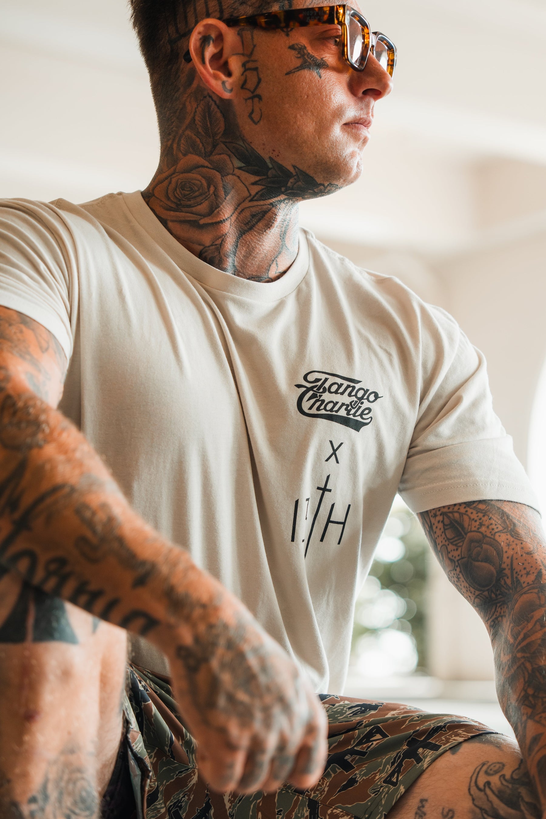 A tattooed man in sunglasses and camouflage shorts wears the Tango Charlie Apparel Lift Heavy. Pray Hard. tee, glancing to the side in a well-lit setting that radiates spiritual resilience.
