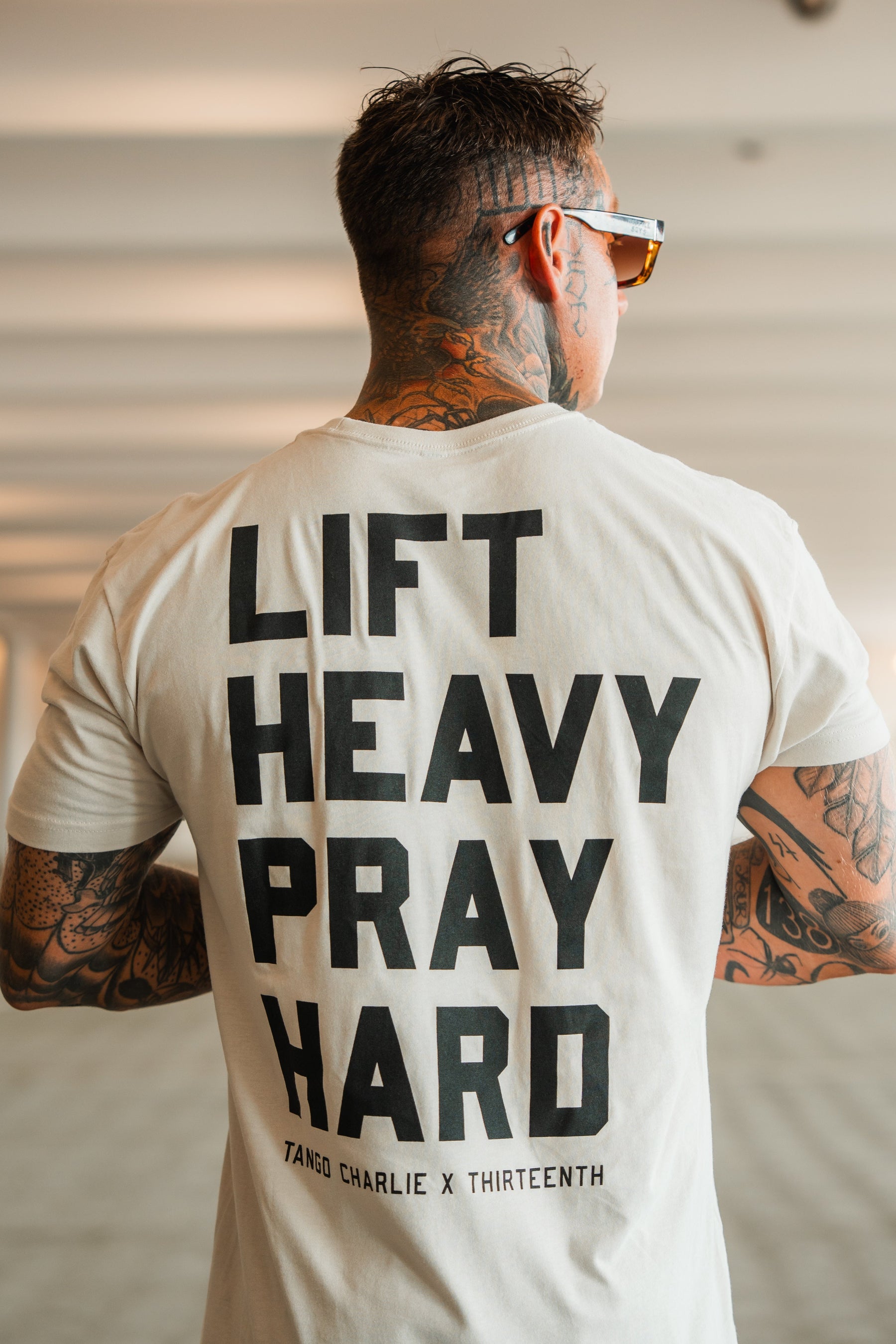 A tattooed man in sunglasses is seen from behind wearing the Lift Heavy. Pray Hard. - Tee by Tango Charlie Apparel, highlighting his spiritual resilience in a blurred indoor setting.