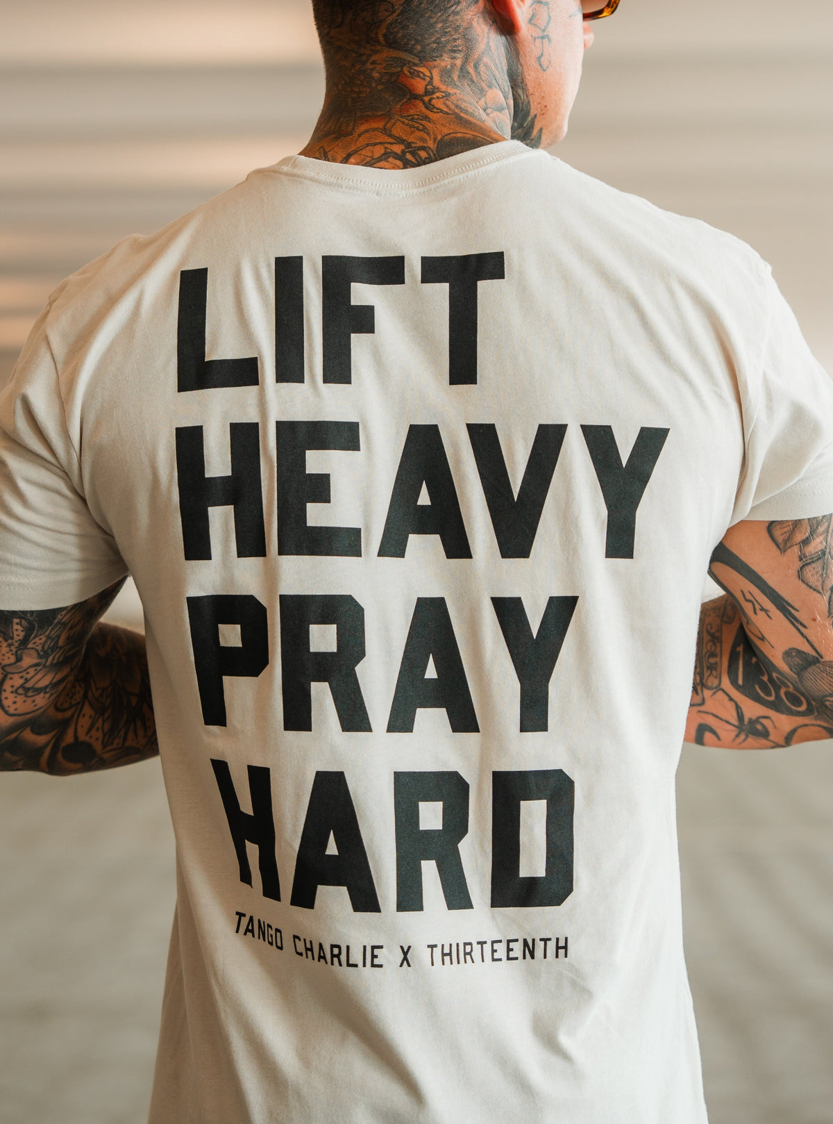 A tattooed person from behind wears a light tee with bold black LIFT HEAVY. PRAY HARD. text, symbolizing resilience. Below is TANGO CHARLIE APPAREL, denoting the brand and product: Lift Heavy. Pray Hard. - Tee by Tango Charlie Apparel.