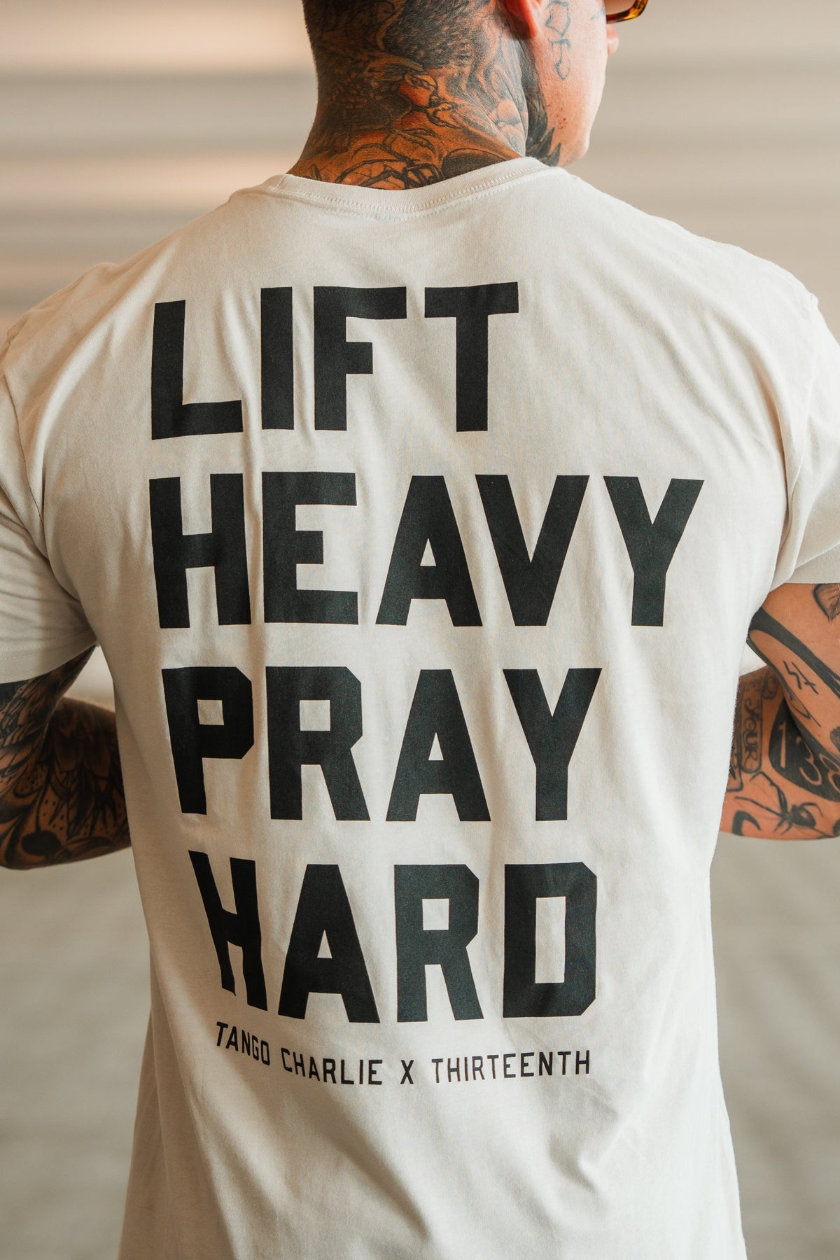 A tattooed person from behind wears a light tee with bold black LIFT HEAVY. PRAY HARD. text, symbolizing resilience. Below is TANGO CHARLIE APPAREL, denoting the brand and product: Lift Heavy. Pray Hard. - Tee by Tango Charlie Apparel.