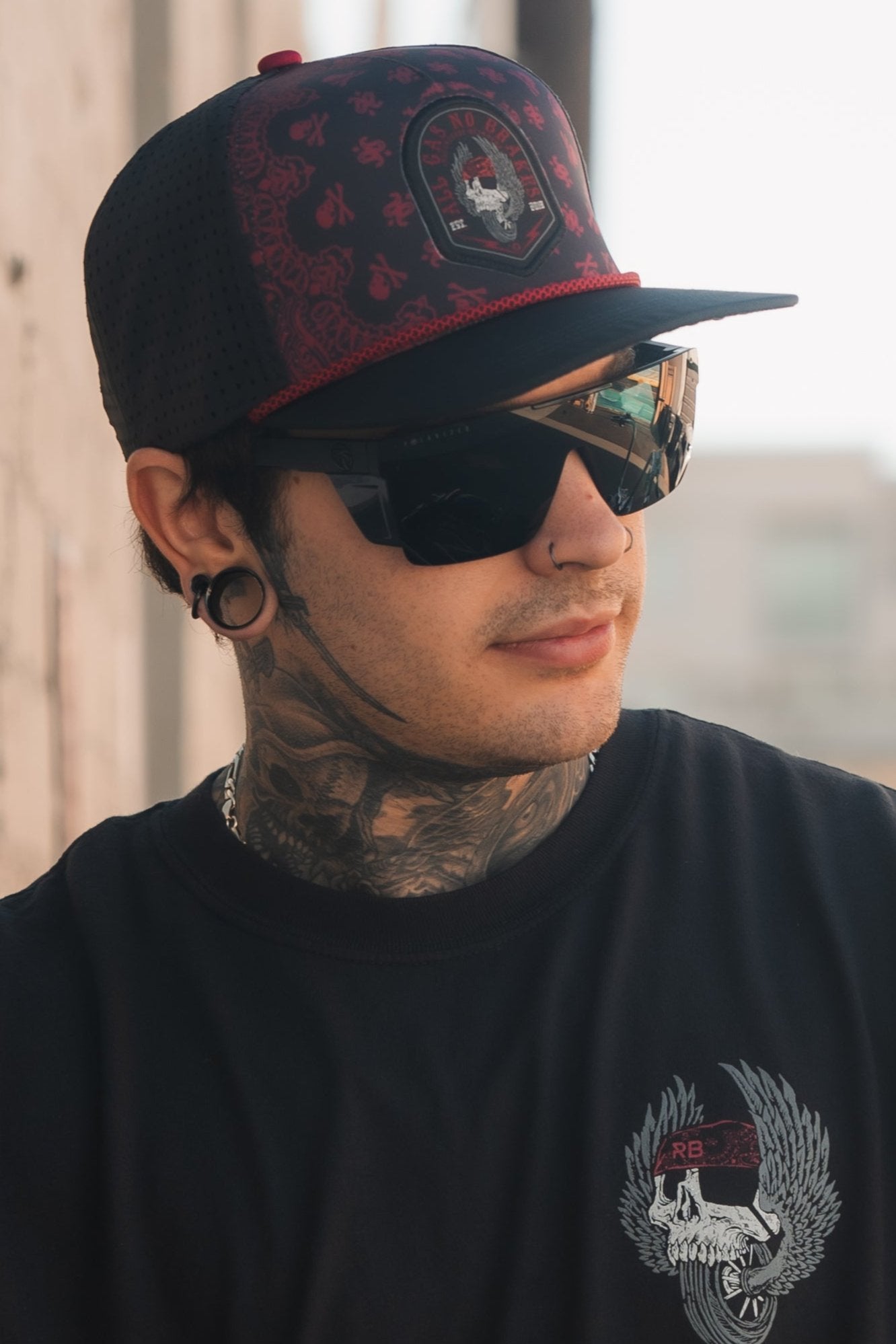 A person wearing an All Gas No Brakes Premium Snapback by Relentless Betrayal, with a skull and crossbones design, dark sunglasses, and a black logo shirt. They have neck tattoos and stand outdoors.