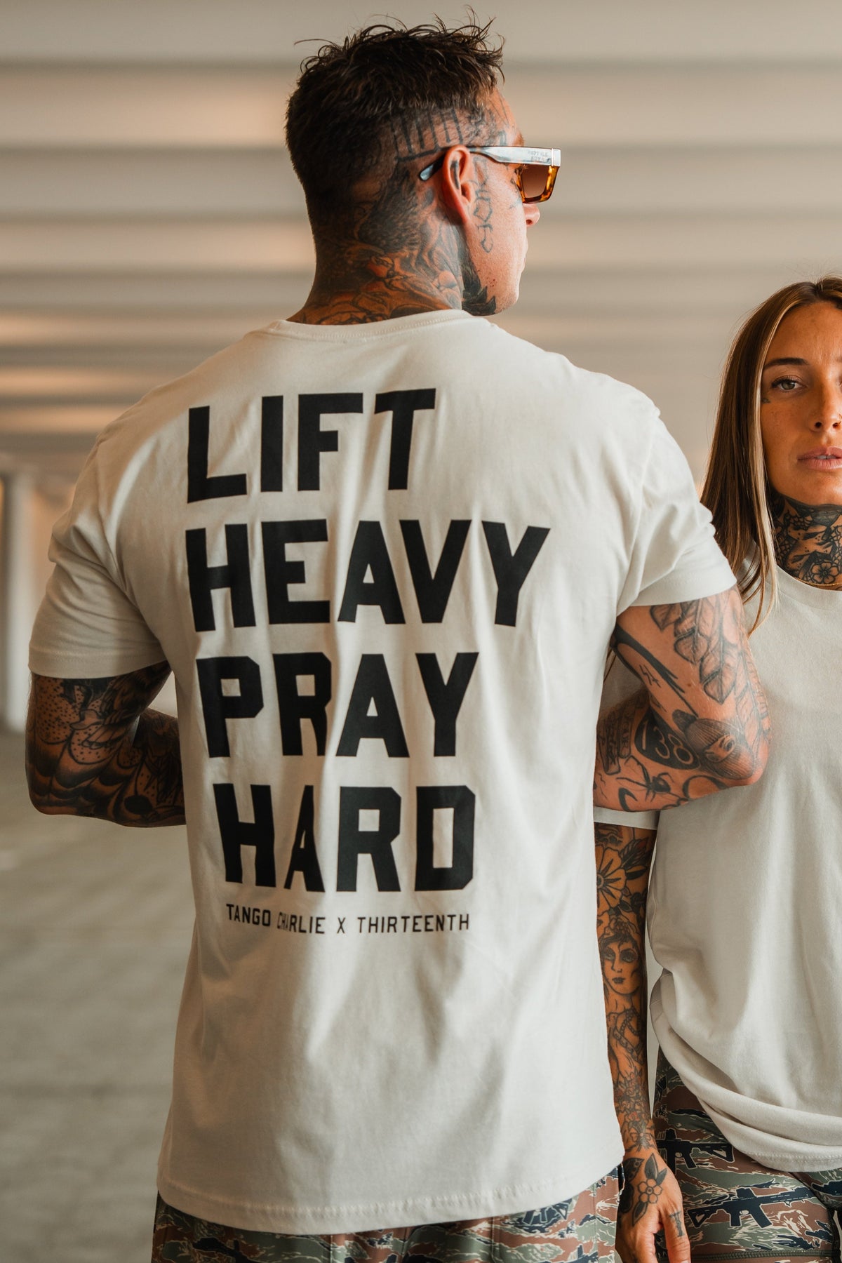 A man with tattoos and sunglasses stands by a woman, wearing the Lift Heavy. Pray Hard. Tee from Tango Charlie Apparel, radiating spiritual resilience in a bright indoor setting.