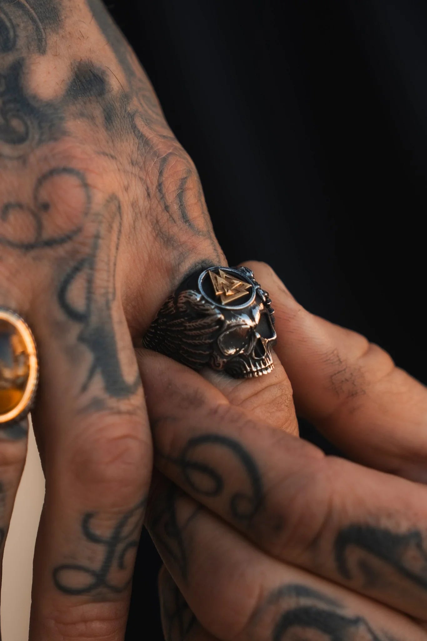 A close-up shows tattooed hands wearing two rings. One flaunts Relentless Betrayals All Gas No Brakes skull ring in stainless steel, adorned with wings and a triangle. The other hand showcases an amber stone ring, with intricate tattoos embodying the swirling essence of youth culture.