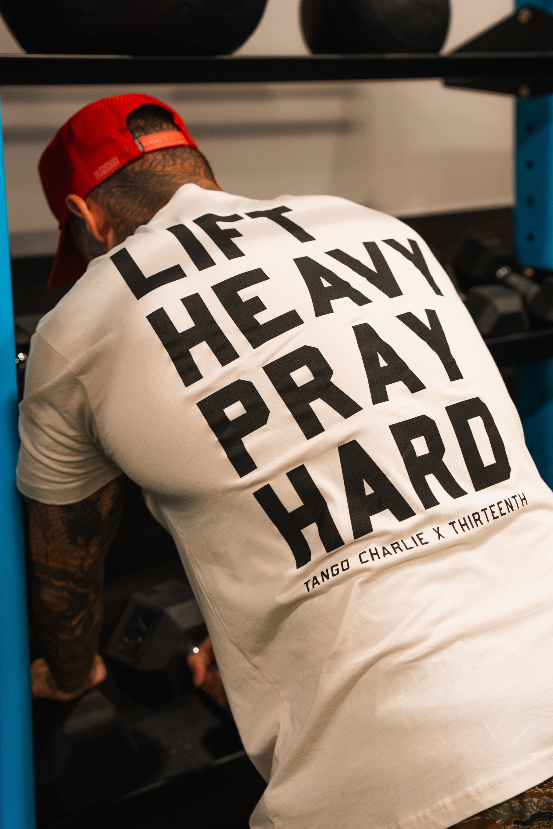 Wearing the Tango Charlie Apparel Lift Heavy. Pray Hard. tee and a red cap, someone leans over gym equipment, ready to lift, representing strength and faith.