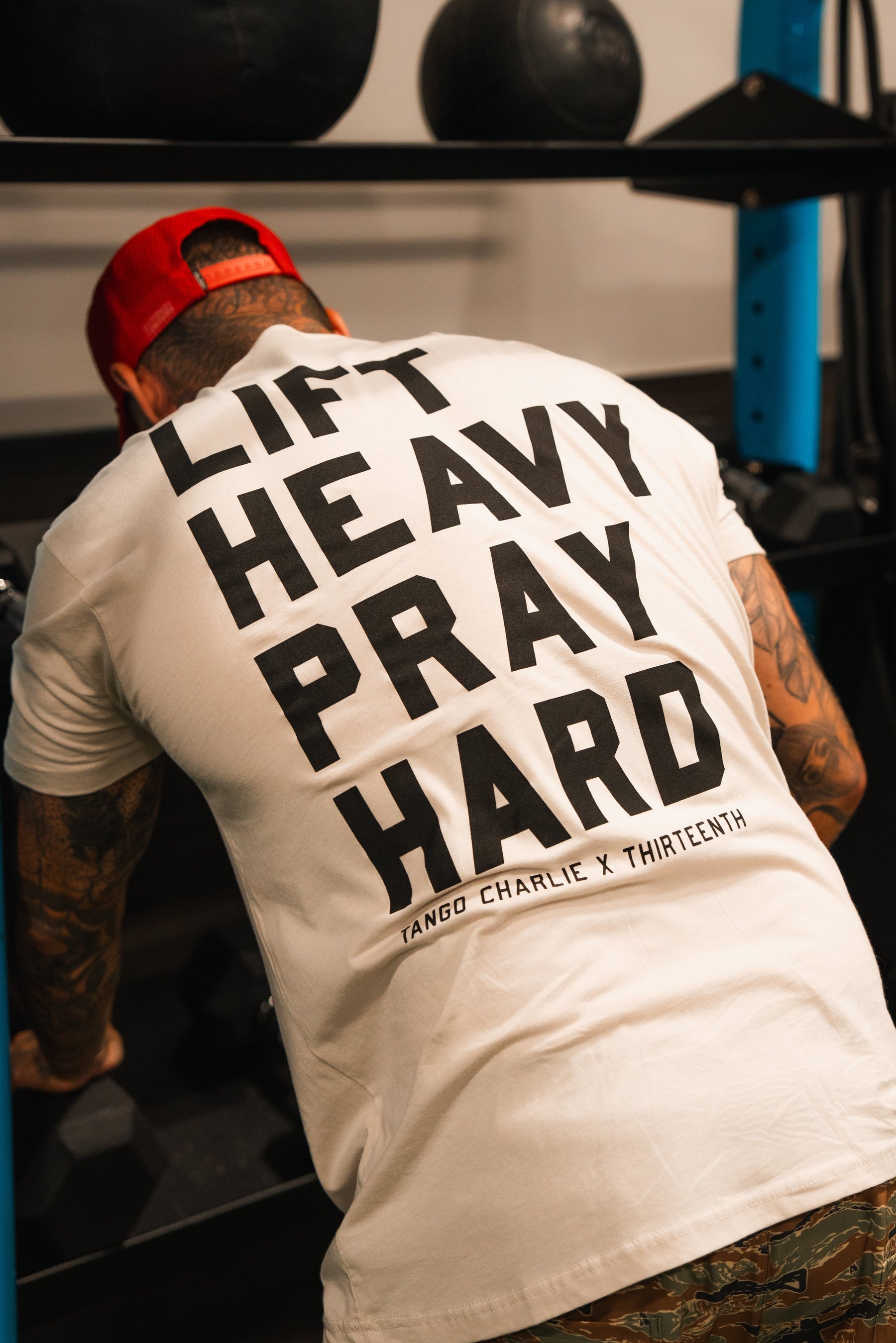A person in a white Lift Heavy. Pray Hard. - Tee by Tango Charlie Apparel and a red cap leans forward in the gym, embodying resilience and faith through strength.