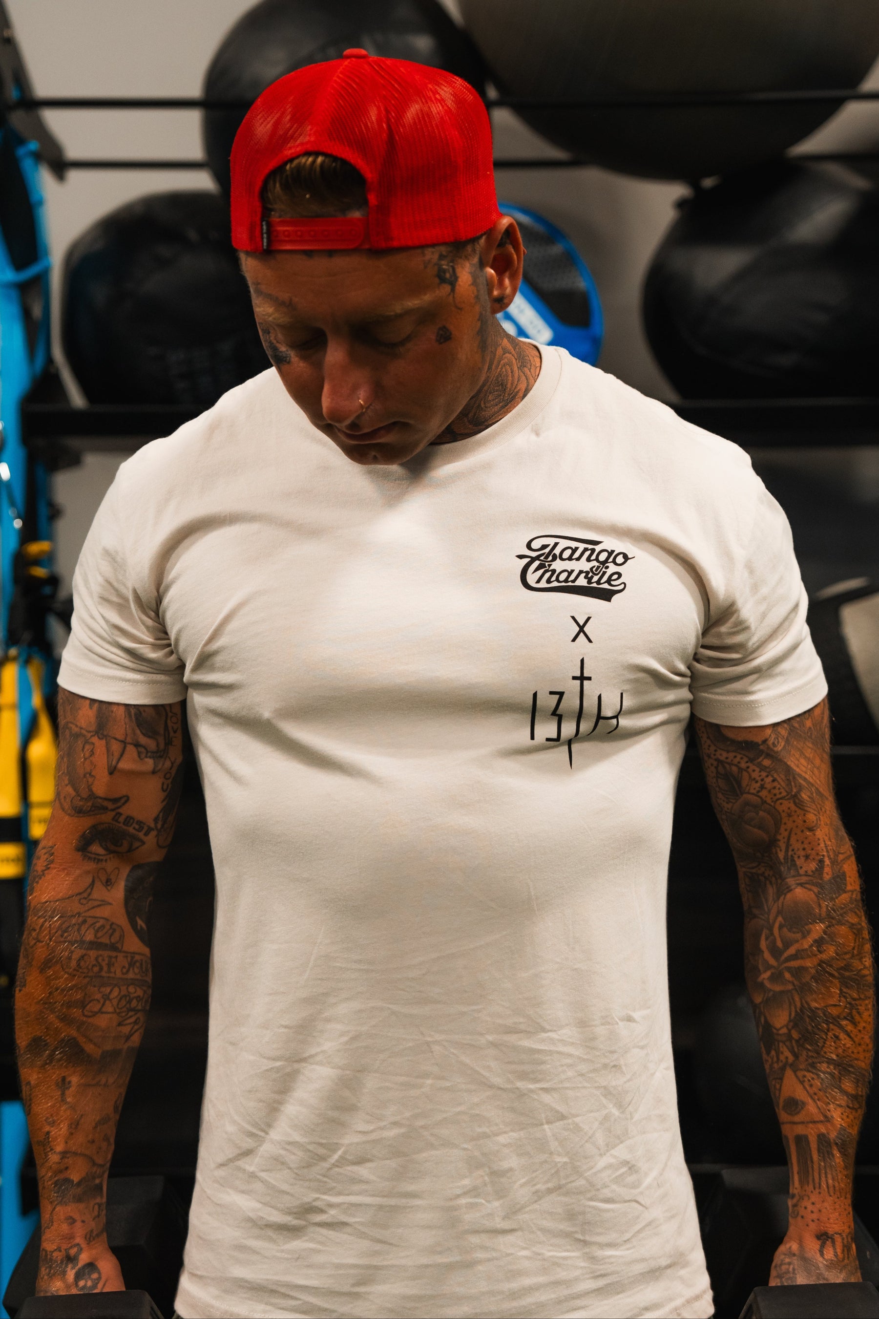 A tattooed man wearing the Lift Heavy. Pray Hard. Tee by Tango Charlie Apparel and a red cap lifts weights in a gym, exuding spiritual resilience amid workout equipment and medicine balls in the background.