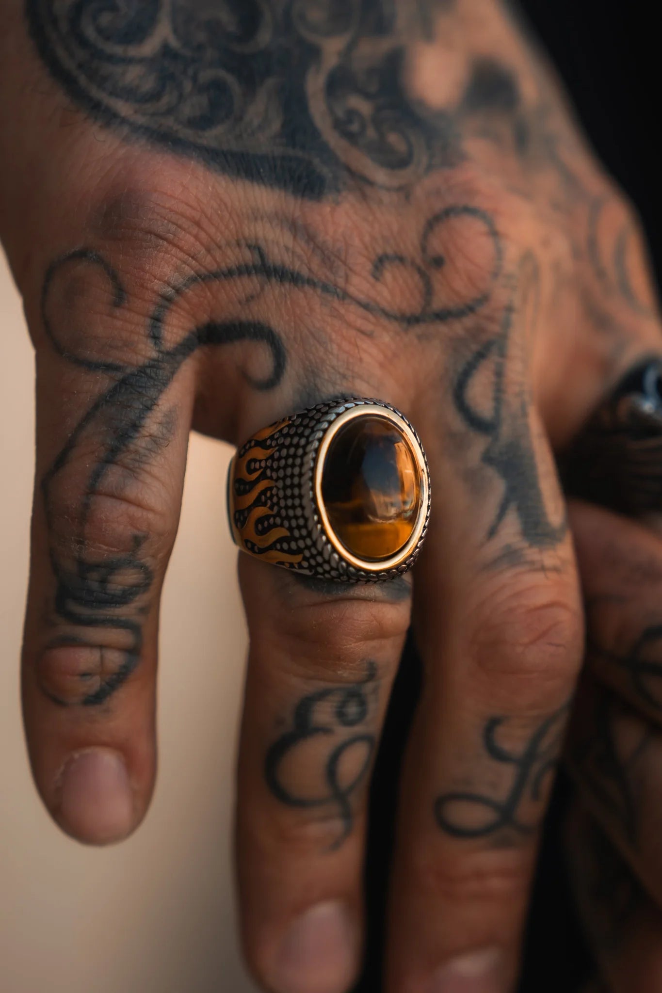 A tattooed hand showcases the handcrafted Inferno Ring by Relentless Betrayal, with its polished amber stone set in intricate stainless steel. Swirling tattoos and letters decorate the skin against a neutral, blurred backdrop.