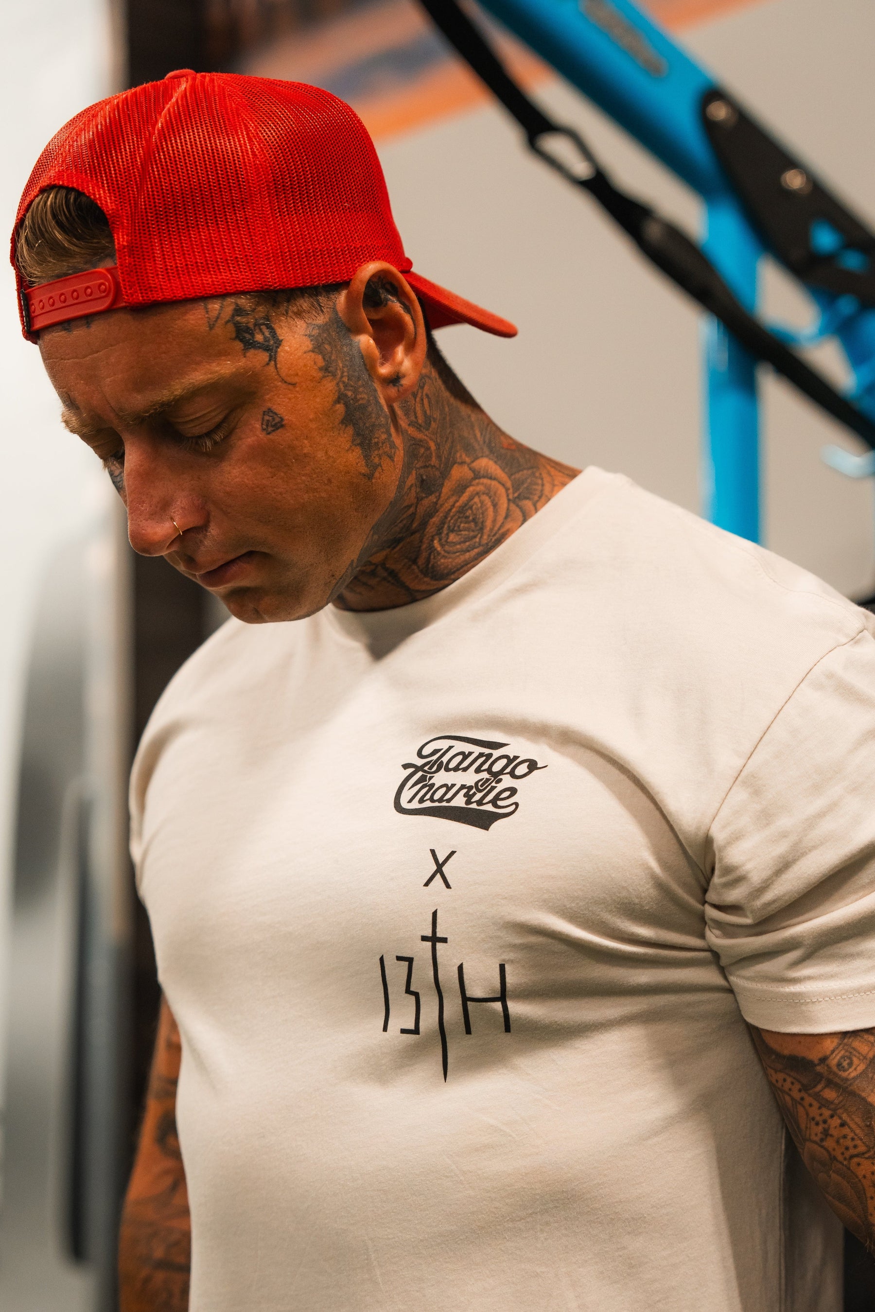 A tattooed man in a red backward cap looks down, wearing the white Lift Heavy. Pray Hard. - Tee by Tango Charlie Apparel, featuring bold black graphics. Blue and black shapes blur in the background.