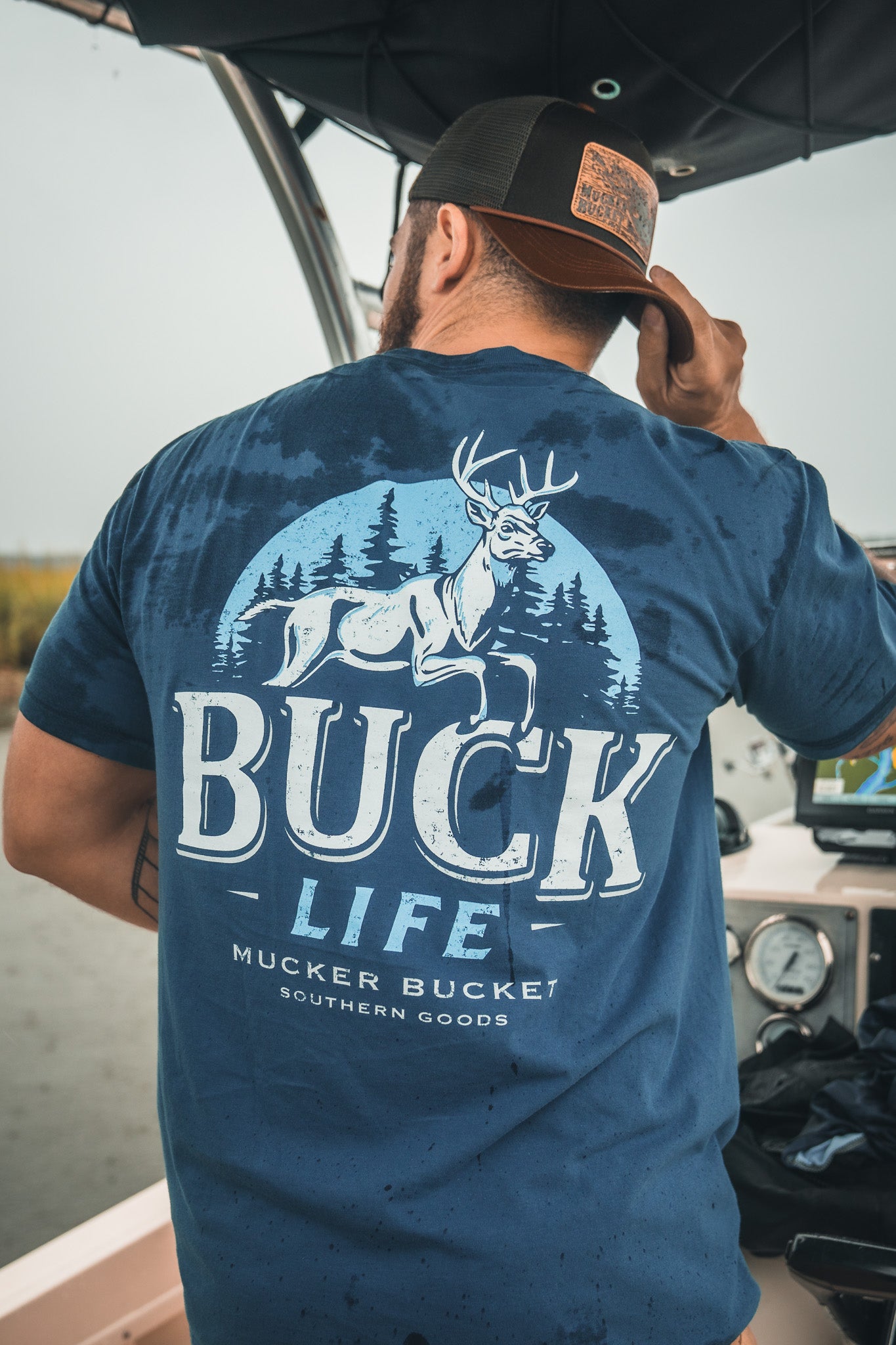 A man in a Mucker Bucket Buck Life Premium Tee, dyed blue and featuring a deer-in-forest graphic, stands on a boat adjusting his cap.