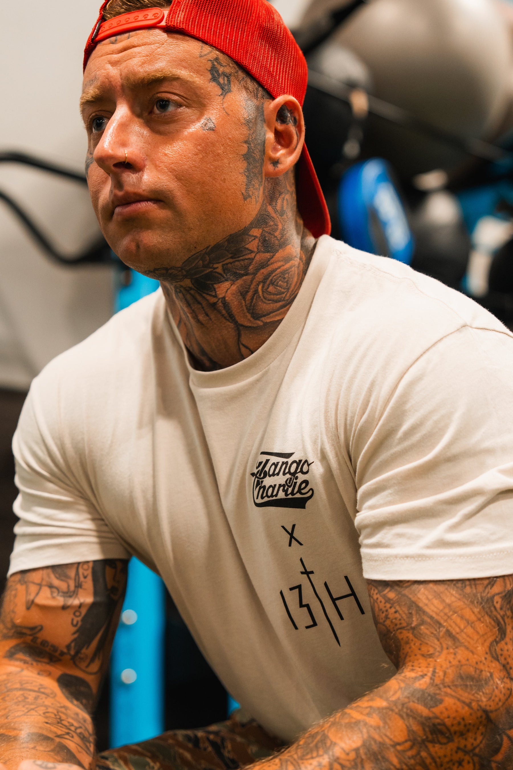 A muscular, tattooed man in a white Lift Heavy. Pray Hard. Tee by Tango Charlie Apparel sits indoors, gym gear behind him, showing determination and embodying the shirtâs motto.