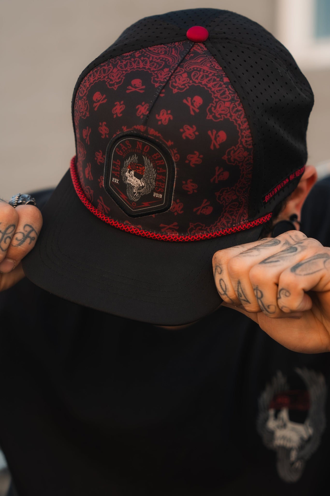 A tattooed person adjusts a black and red All Gas No Brakes Premium Snapback by Relentless Betrayal, featuring bandana print and skull logo, while wearing a matching black shirt with the same skull design. Their face is hidden.