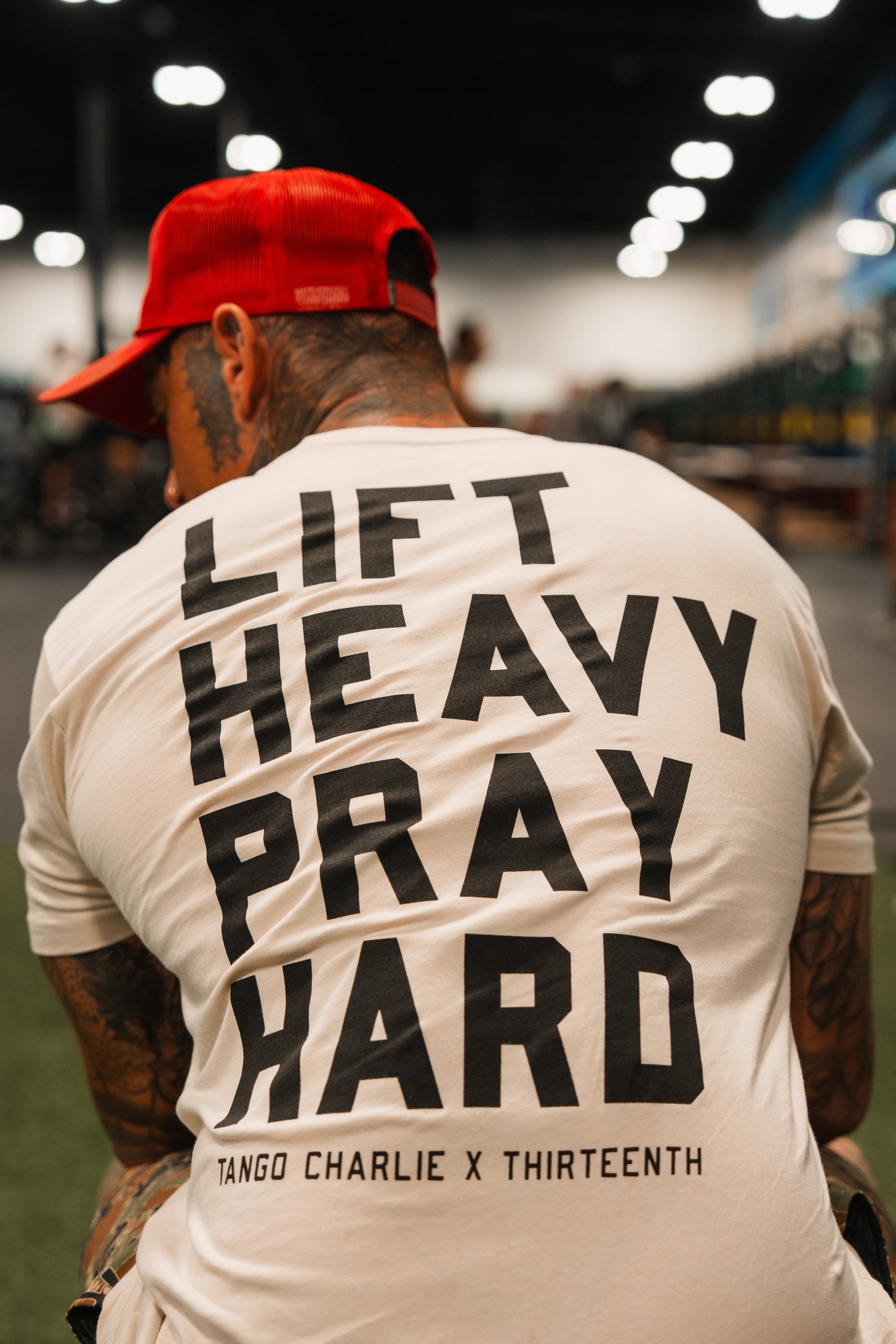 Seen from behind in a gym, a person wears the Lift Heavy. Pray Hard. Tee by Tango Charlie Apparelâa white shirt with bold black textâexemplifying spiritual resilience and dedication.