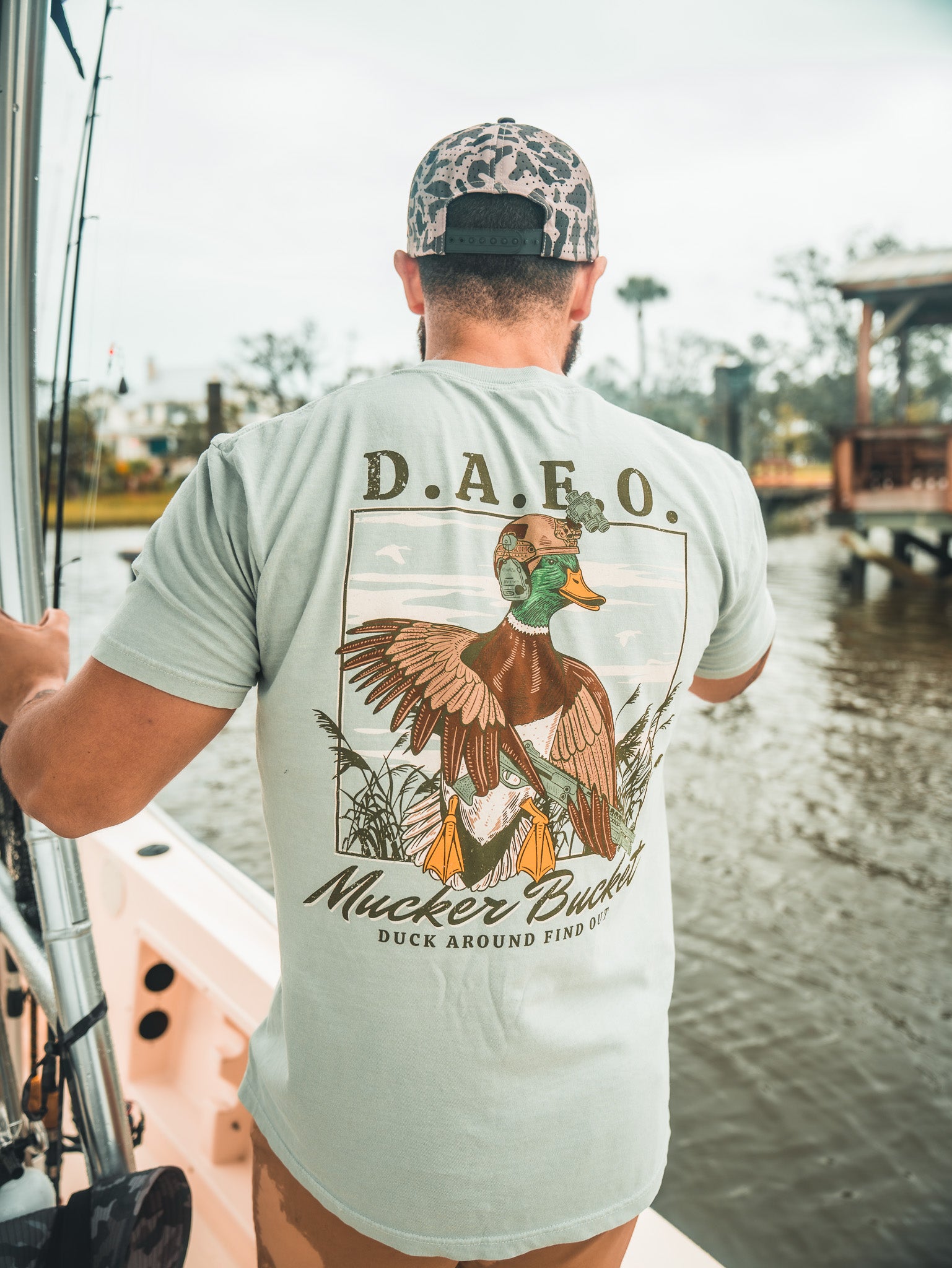 A man on a boat holds a fishing rod while wearing the DAFO Premium Tee by Mucker Bucket, featuring an illustrated duck and Duck Around Find Out. Water and trees can be seen in the background.