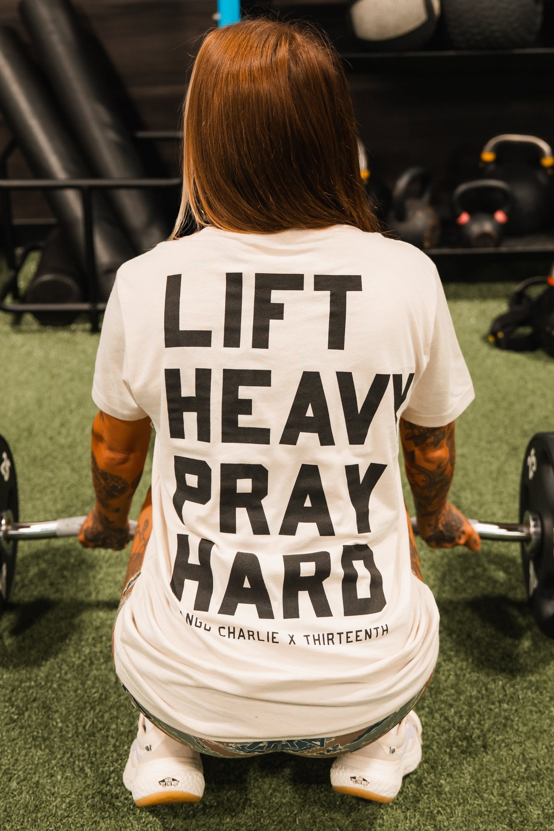 A tattooed individual squats with a barbell, wearing the Lift Heavy. Pray Hard. Tee by Tango Charlie Apparel. The T-shirtâs bold slogan reflects strength and faith, with gym equipment visible in the background.