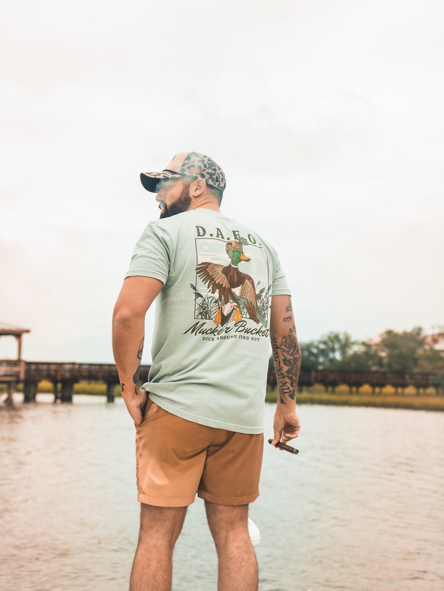 A man in a cap, Mucker Bucket DAFO Premium Tee, and shorts stands by the water holding a cigar, facing away with trees, a dock, and cloudy sky in the background.
