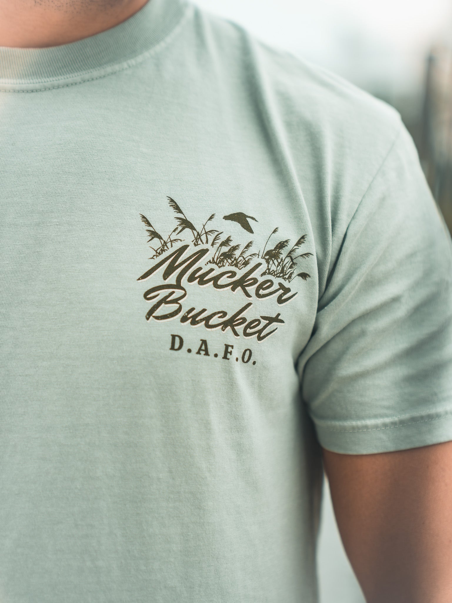 A person wearing the Mucker Bucket DAFO Premium Tee, a relaxed fit light green shirt with a garment-dyed finish and a palm tree, bird, and “Mucker Bucket D.A.F.O.” graphic on the left chest.