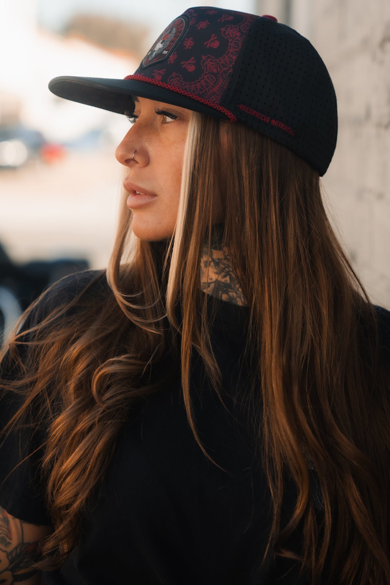A person with long hair dons the black and red All Gas No Brakes Premium Snapback by Relentless Betrayal, subtly adorned with a skull and crossbones. Paired with a black shirt, they gaze sideways, revealing neck tattoos against a softly blurred background.