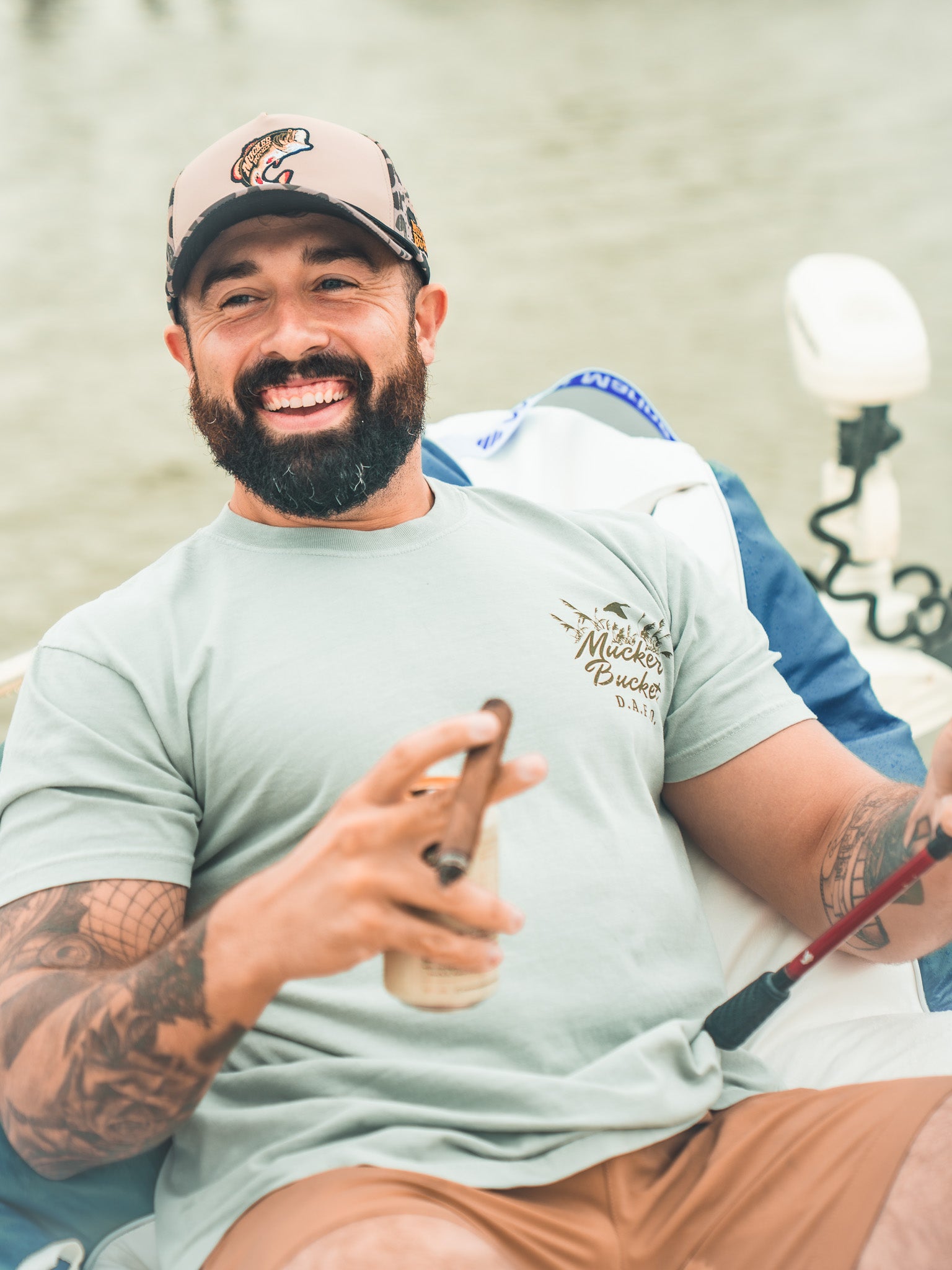 A smiling bearded man with tattoos relaxes on a boat, holding a fishing rod and cigar. He wears the Mucker Bucket DAFO Premium Tee in light blue and a beige cap with fish logo, with water in the background.
