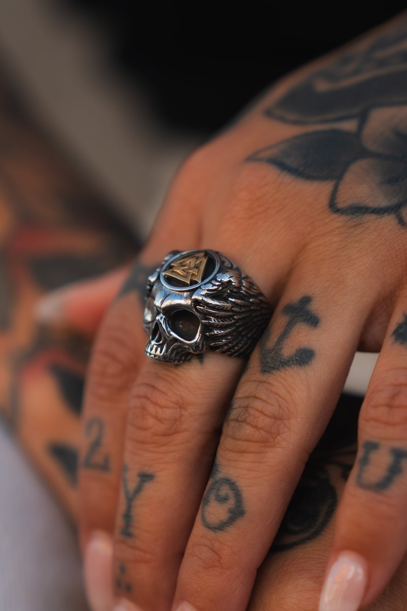Close-up of a tattooed hand wearing the All Gas No Brakes Ring by Relentless Betrayal, a stainless steel skull with wings, capturing youth culture. Numbers and designs adorn the index and middle fingers, while the blurred background enhances the rings rebellious aura.