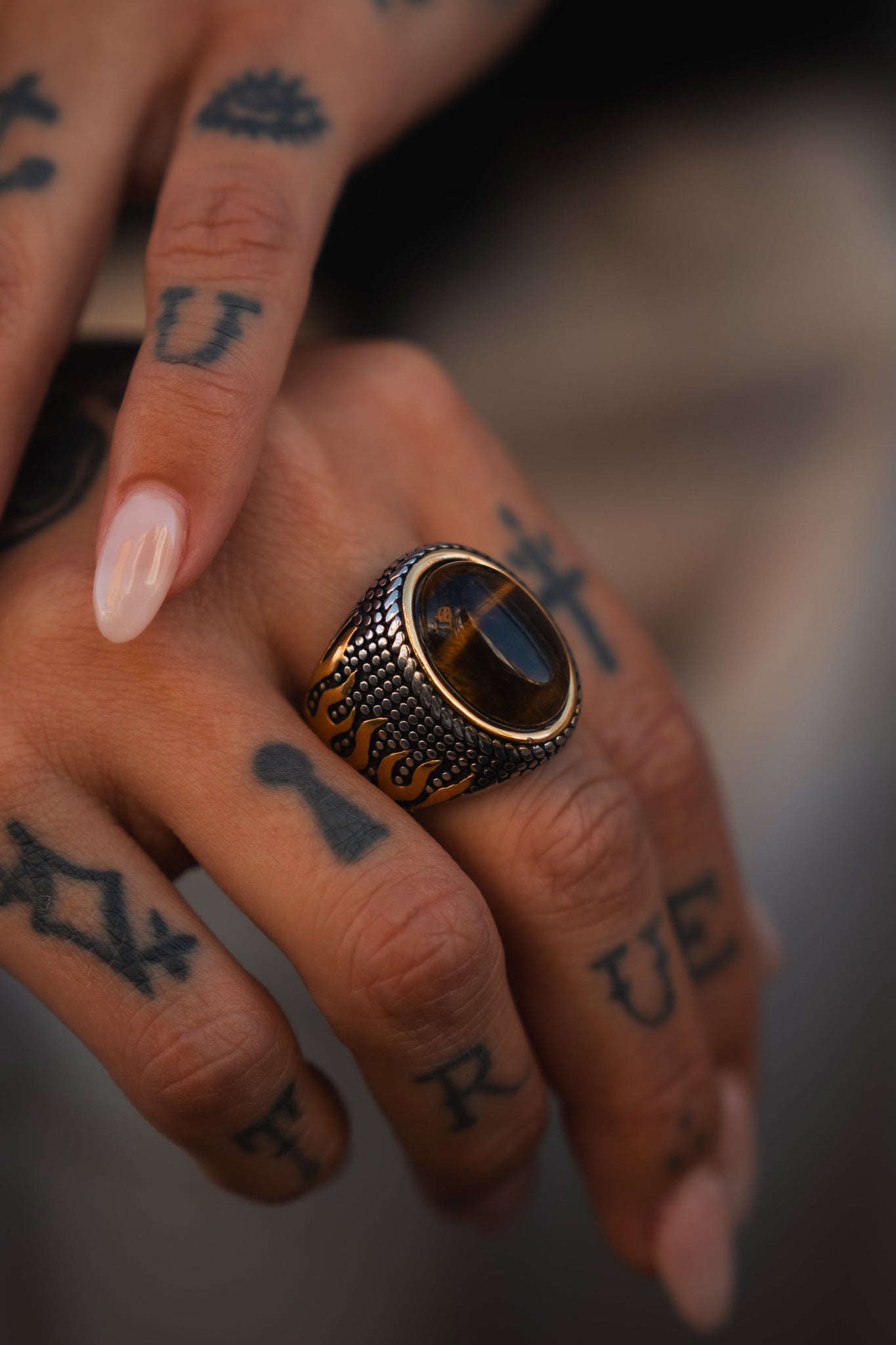 A close-up of a tattooed hand with TRUE on the fingers showcases the Relentless Betrayals handcrafted Inferno Ring, featuring a dark stone. The polished, nude nails provide an elegant contrast to this stunning stainless steel piece.
