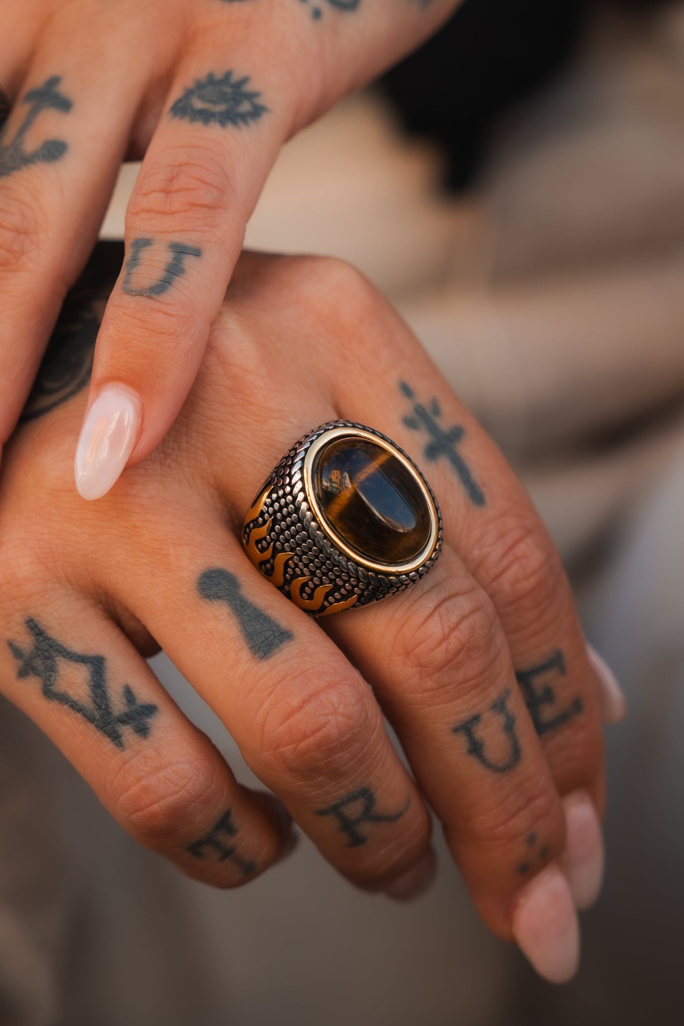 A tattooed hand with manicured natural nails showcases the handcrafted Inferno Ring by Relentless Betrayal, featuring a dark stone and dazzling stainless steel accents.