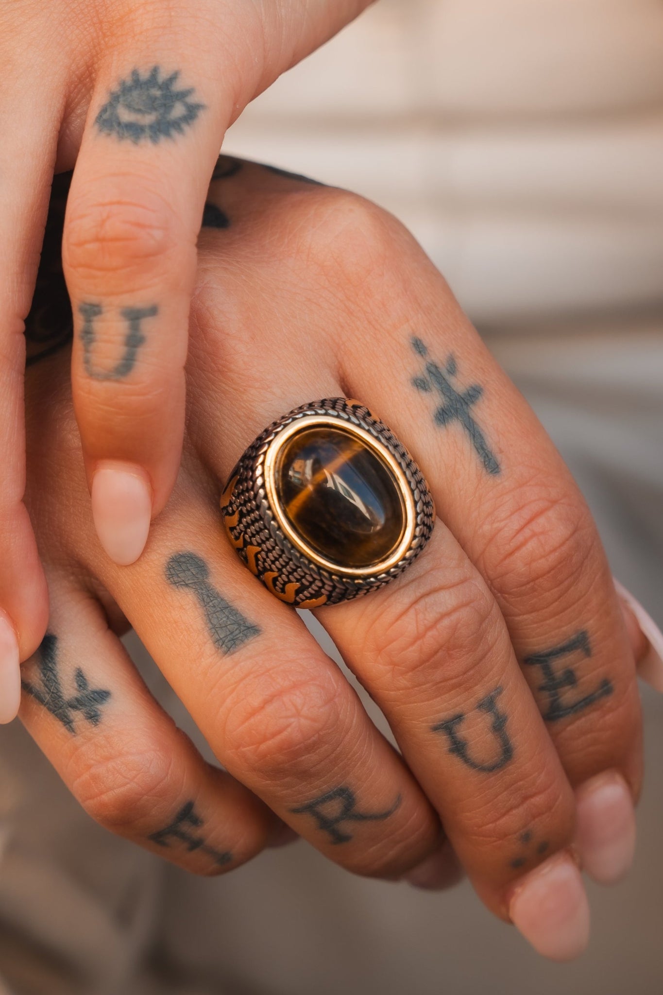 A tattooed hand holds another, highlighting an ornate Inferno Ring by Relentless Betrayal. The dark, reflective stone on the ring finger contrasts with the inked letters and symbols on the skin against a light background.