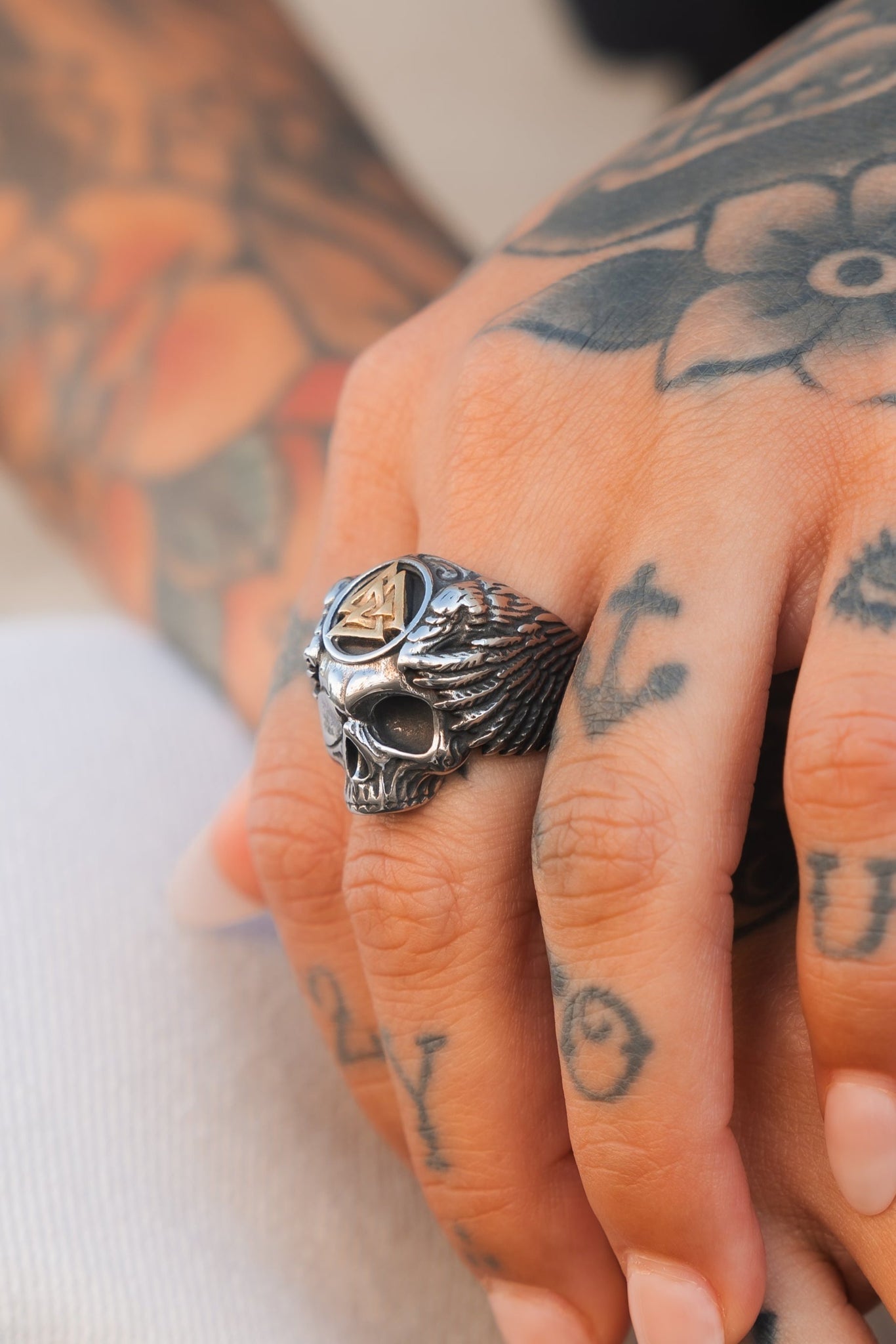 A tattooed hand adorned with the Relentless Betrayals All Gas No Brakes Ring, a stainless steel skull with wing design, rests on a surface. Vivid tattoos of anchors, numbers, and a flower capture the essence of youthful culture.