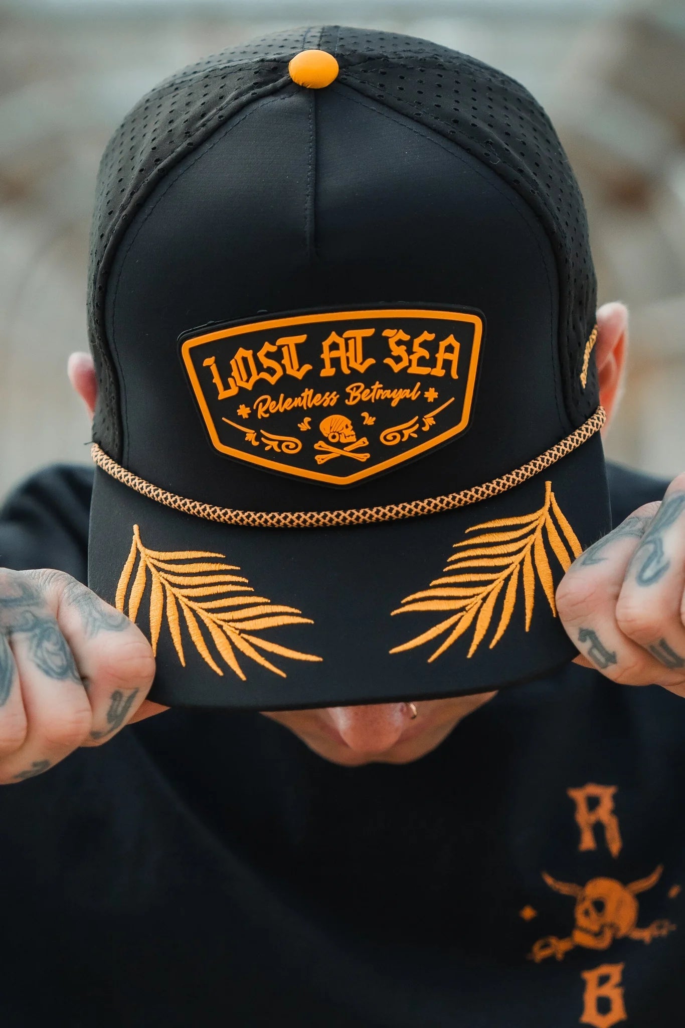 Wearing the Lost At Sea PVC Snapback by Relentless Betrayal, a person sports a black cap with vibrant orange Lost at Sea text. The brim features orange palm leaves, while tattooed hands grip the cap and a lip piercing adds to their pirate vibe.