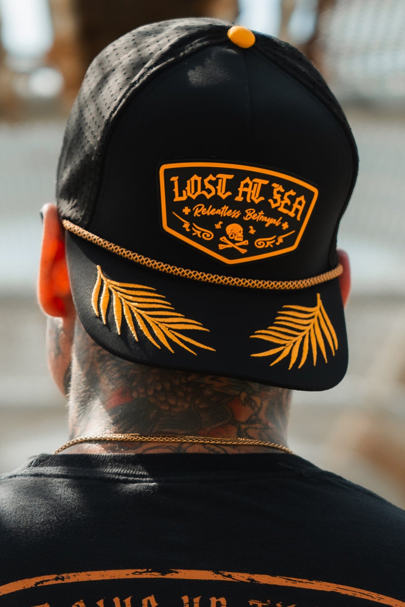 A neck-tattooed person wears the Relentless Betrayal Lost At Sea PVC Snapback, featuring yellow embroidery of a skull and palm fronds. Perfect for pirate enthusiasts, it stands out against a blurred background.