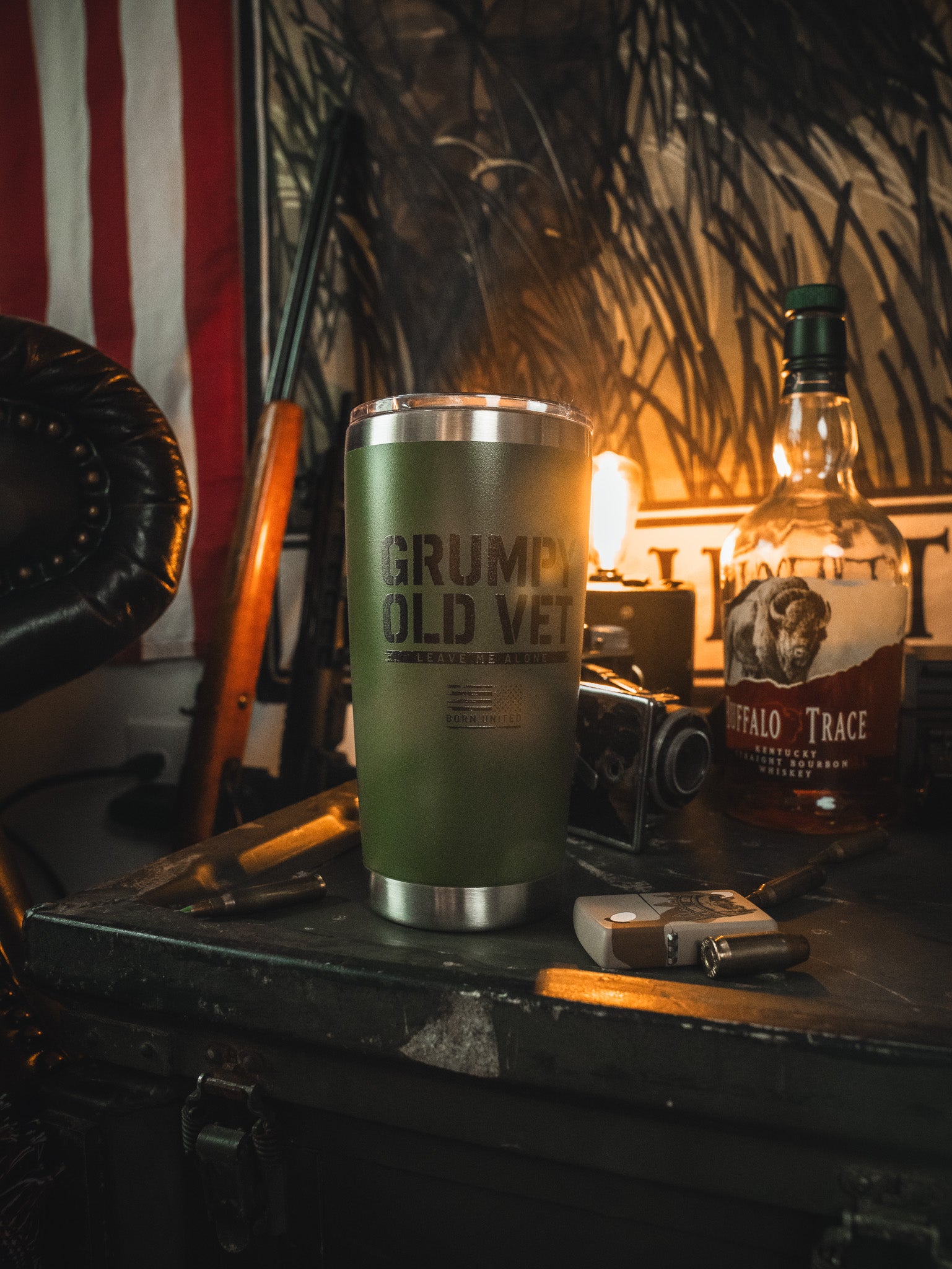 The Born United Private Label Grumpy Old Vet Tumbler - 20oz sits on a cluttered table with a whiskey bottle, camera, bullets, and lighter, as a warm lamp highlights an American flag and rifle in the background.