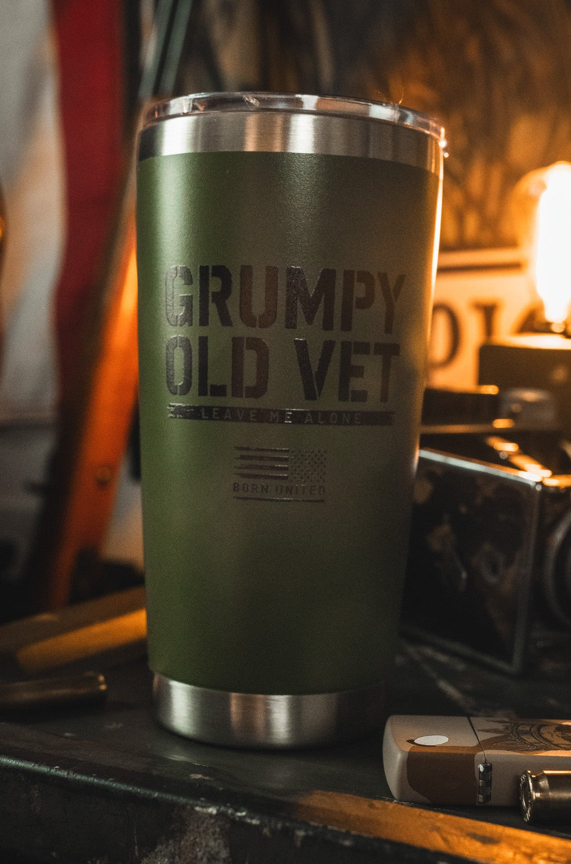 A Born United Private Label 20oz Grumpy Old Vet Tumbler in green, featuring the printed phrase, rests among vintage items on a cluttered surface with a glowing bulb in the background.