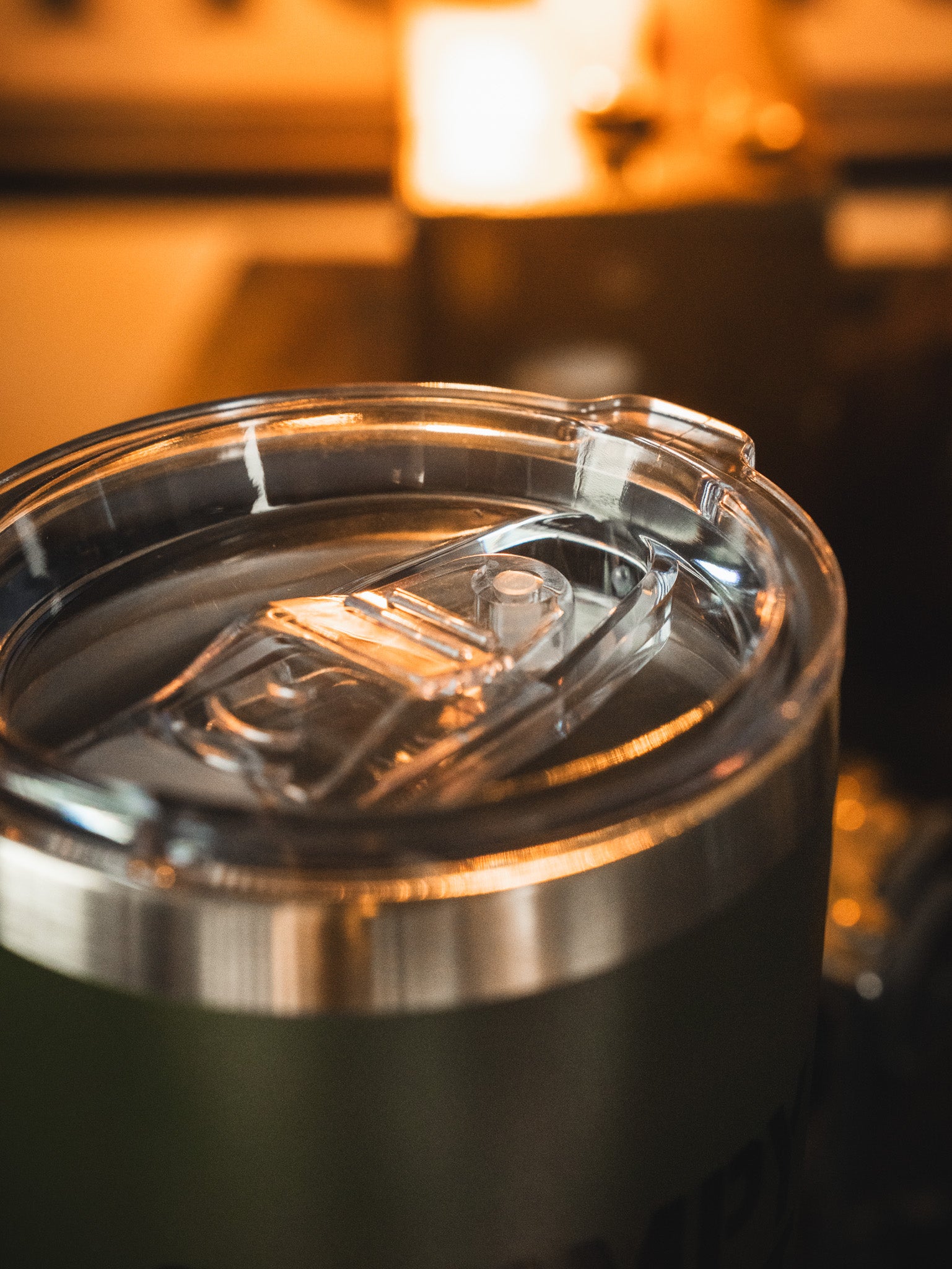 The Grumpy Old Vet Tumbler - 20oz by Born United Private Label features a sleek stainless steel design with a clear lid, filled with a beverage and set against a warmly lit, softly blurred background.