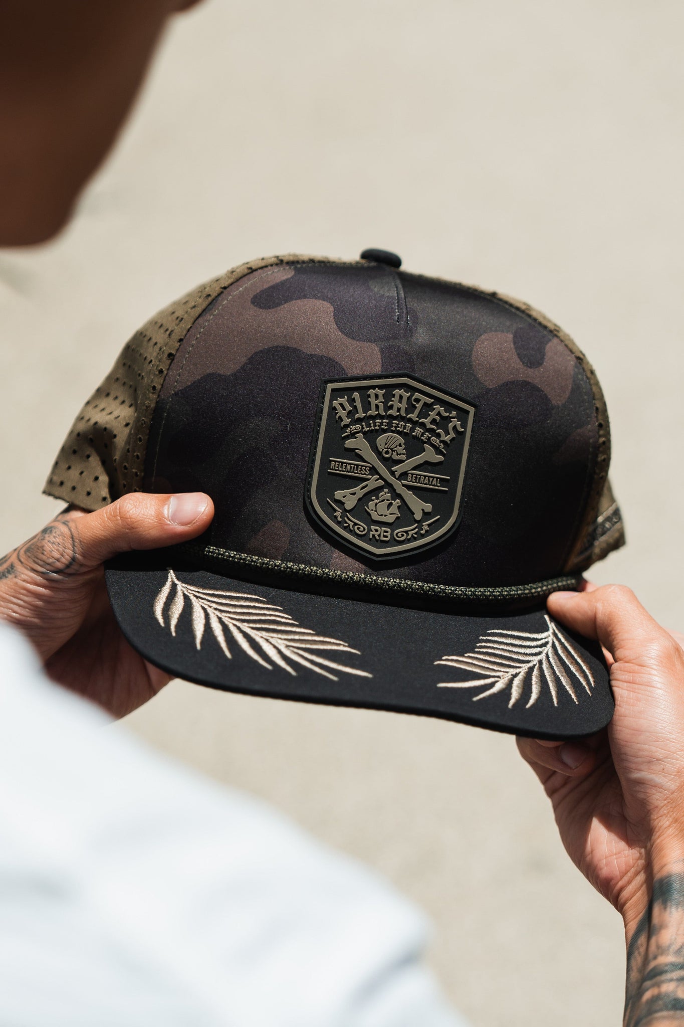 A person holds the Pirates Life Camo Snapback by Relentless Betrayal, a rugged camo baseball cap with crossed anchors and Pirate text patch, featuring perforated sides and embroidered palm leaves on the brim, capturing the adventurous spirit of a pirates life.