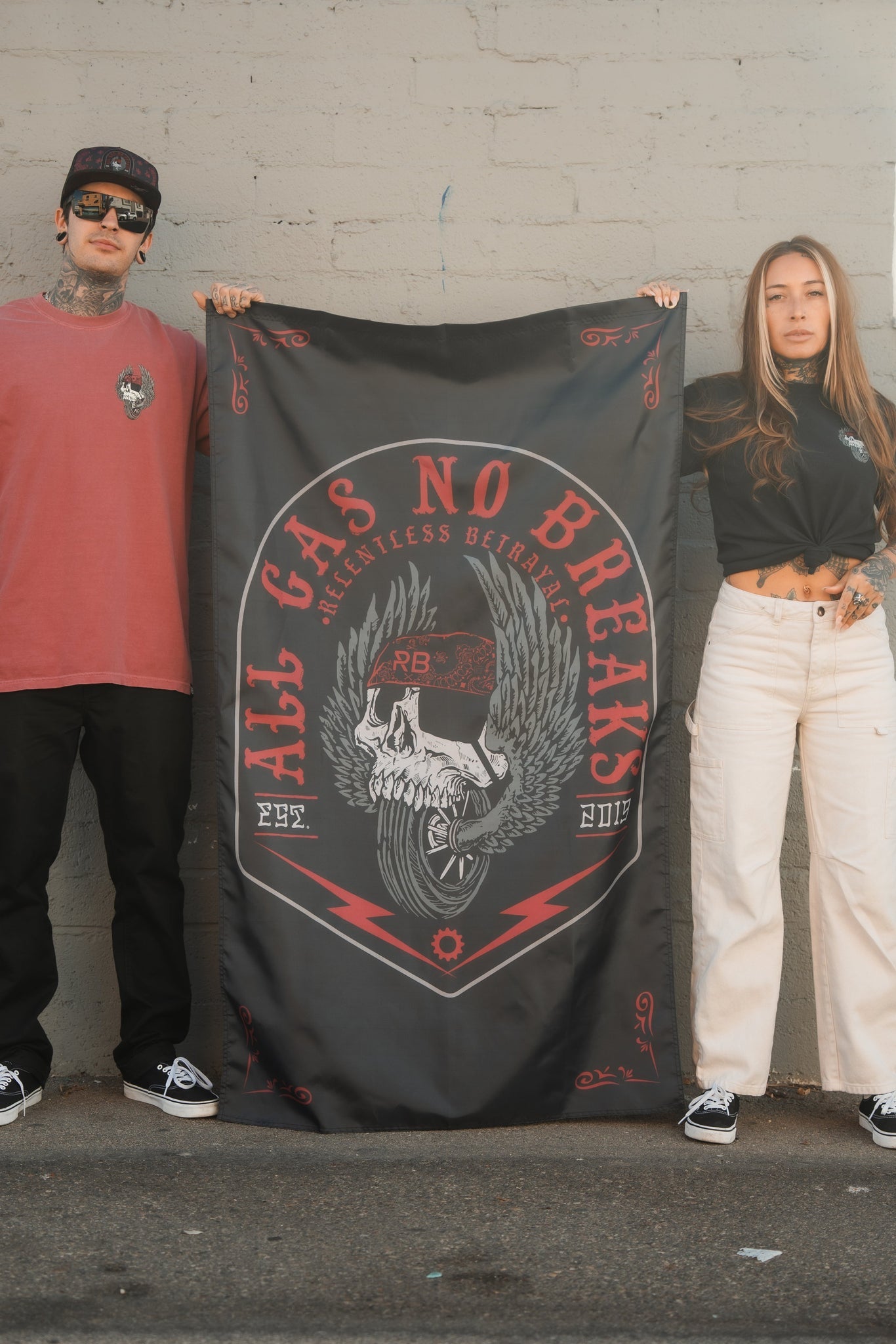 Two people pose in front of a brick wall, showcasing the youthful vibe of Relentless Betrayals All Gas No Brakes Flag, featuring a skull with wings and a motorcycle wheel. The design includes red lightning motifs and the text All Gas No Break, echoing energy and drive. Established in 2017.