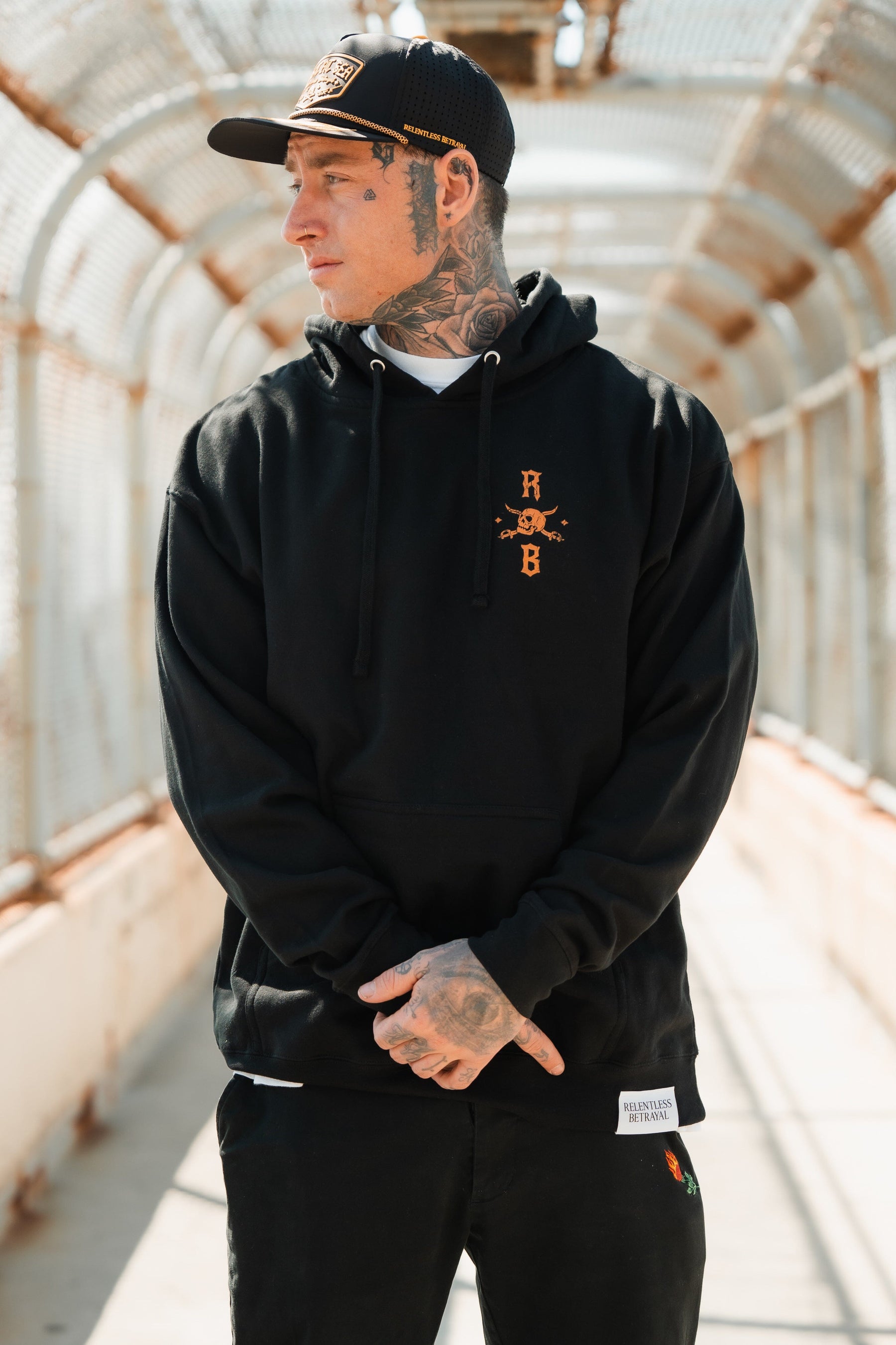 With tattoos on neck and face, a person stands on a pedestrian bridge in a black Standfast Gold Foil Hoodie from Relentless Betrayal, crafted from a comfy cotton/polyester blend. Amid the metal mesh, they gaze sideways with clasped hands, embodying unisex style.