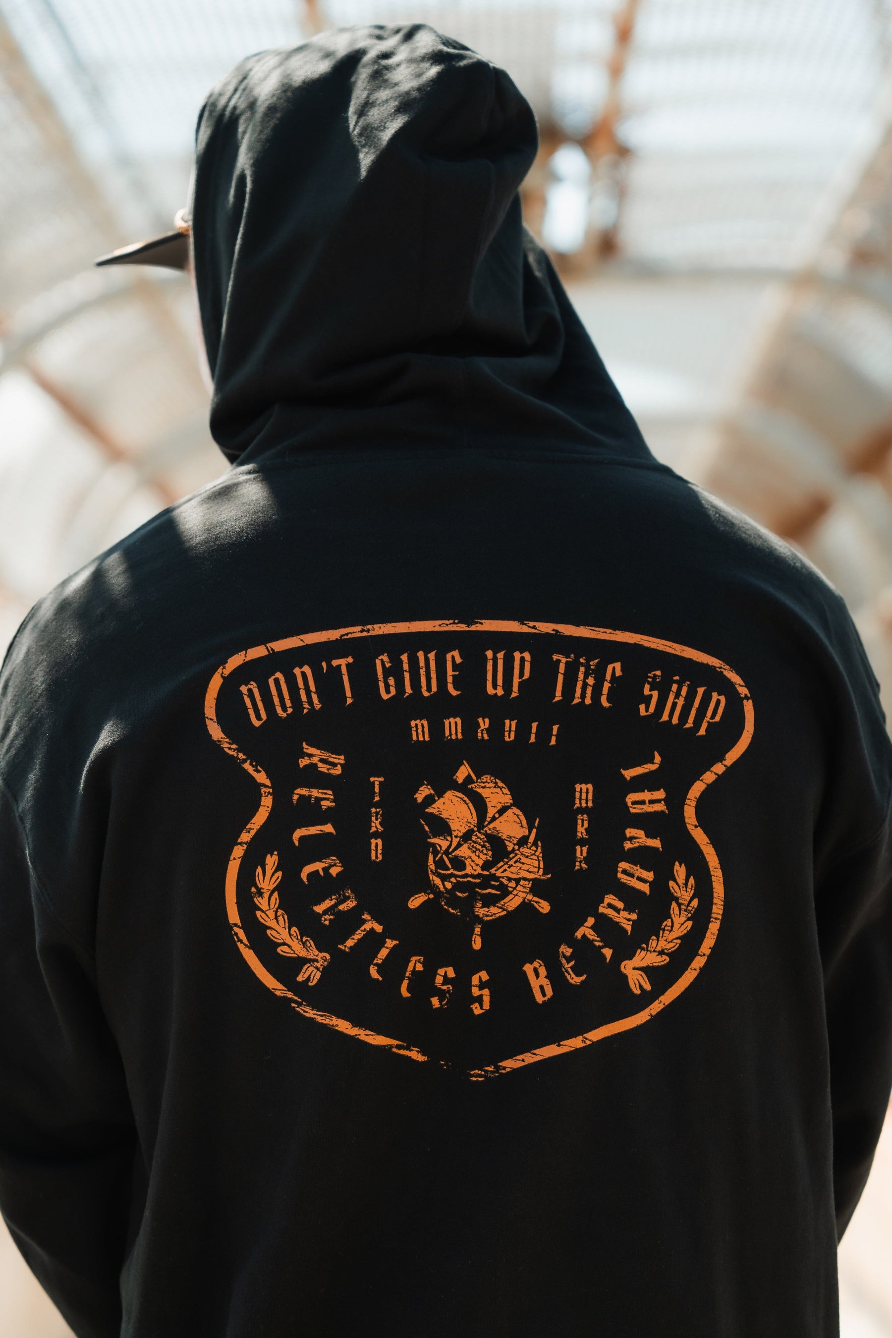 In a covered walkway, someone wears a Standfast Gold Foil Hoodie by Relentless Betrayal. Its unisex design highlights a back graphic of a ship with Dont Give Up The Ship and Relentless Betrayal, crafted from a comfy cotton/polyester blend, mirroring the resilience of its message.
