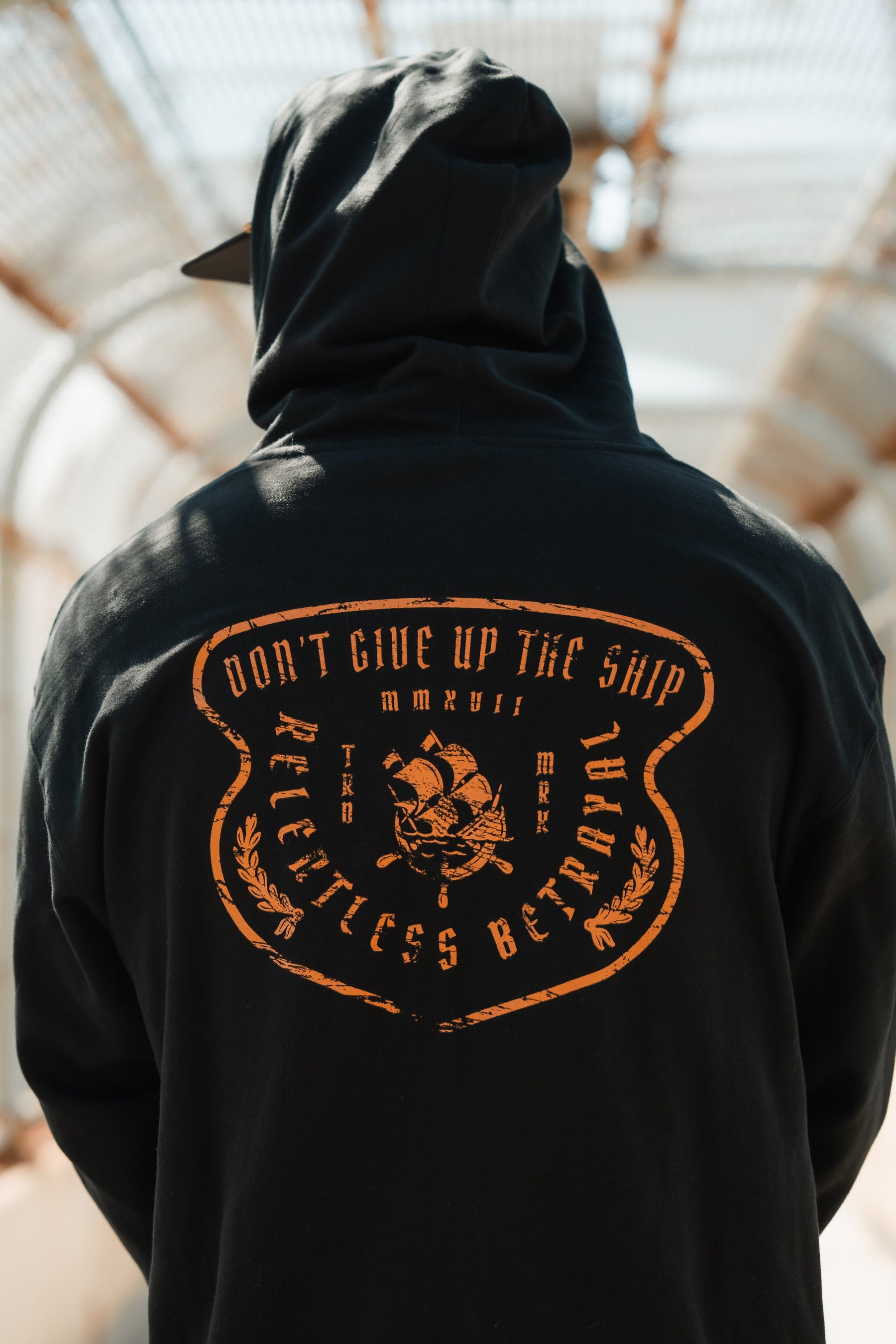 A person wears a Relentless Betrayal unisex Standfast Gold Foil Hoodie made from a cotton/polyester blend. It features an orange shield with DONT GIVE UP THE SHIP and RELENTLESS BETRAYAL. A ship design is central to the look, and the hood stylishly conceals their face.