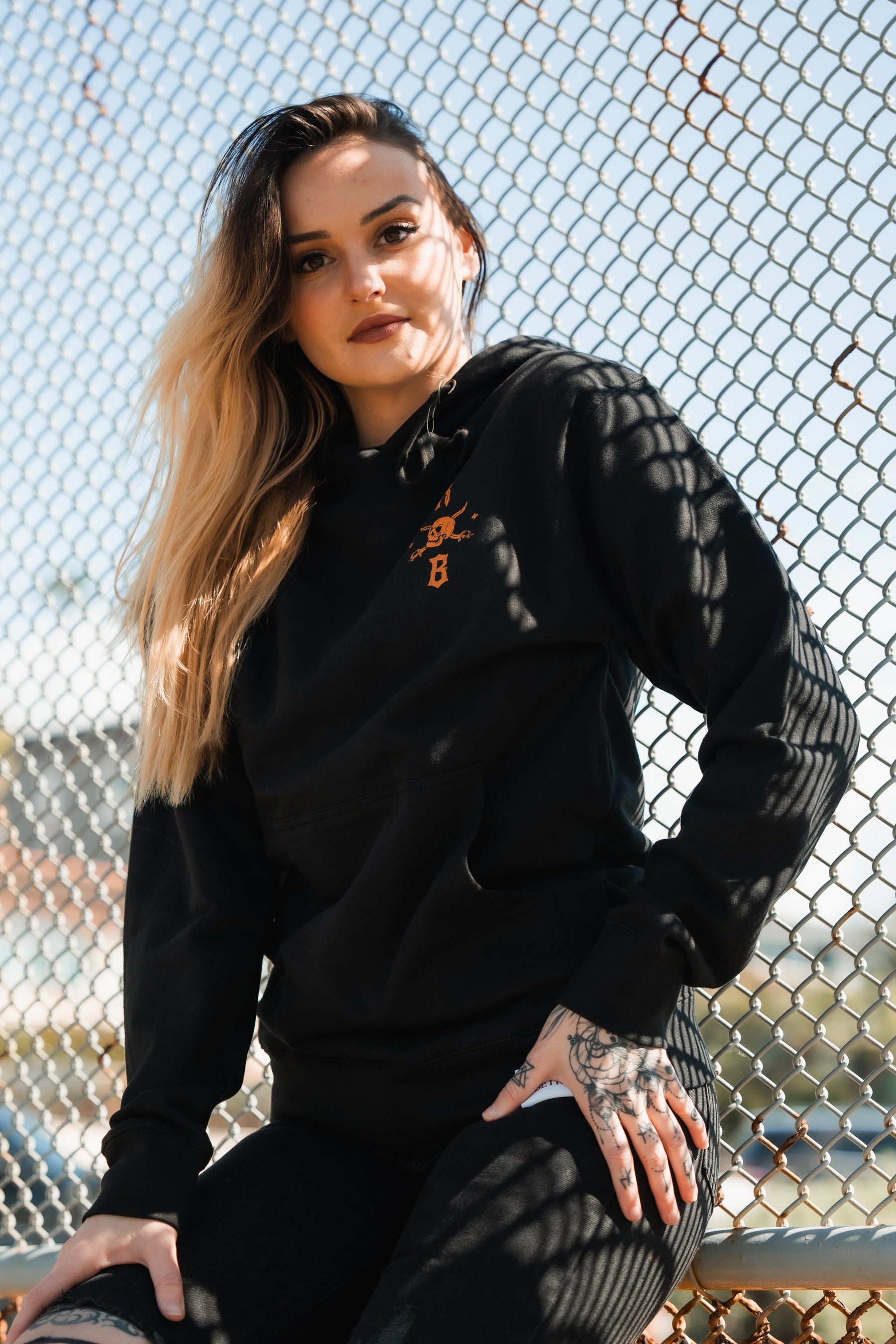 A long-haired person wearing a Standfast Gold Foil Hoodie and pants by Relentless Betrayal sits against a chain-link fence. Sunlight casts shadows on their face and clothing. One tattooed hand rests on their lap. This unisex hoodie is made from a comfortable cotton/polyester blend.