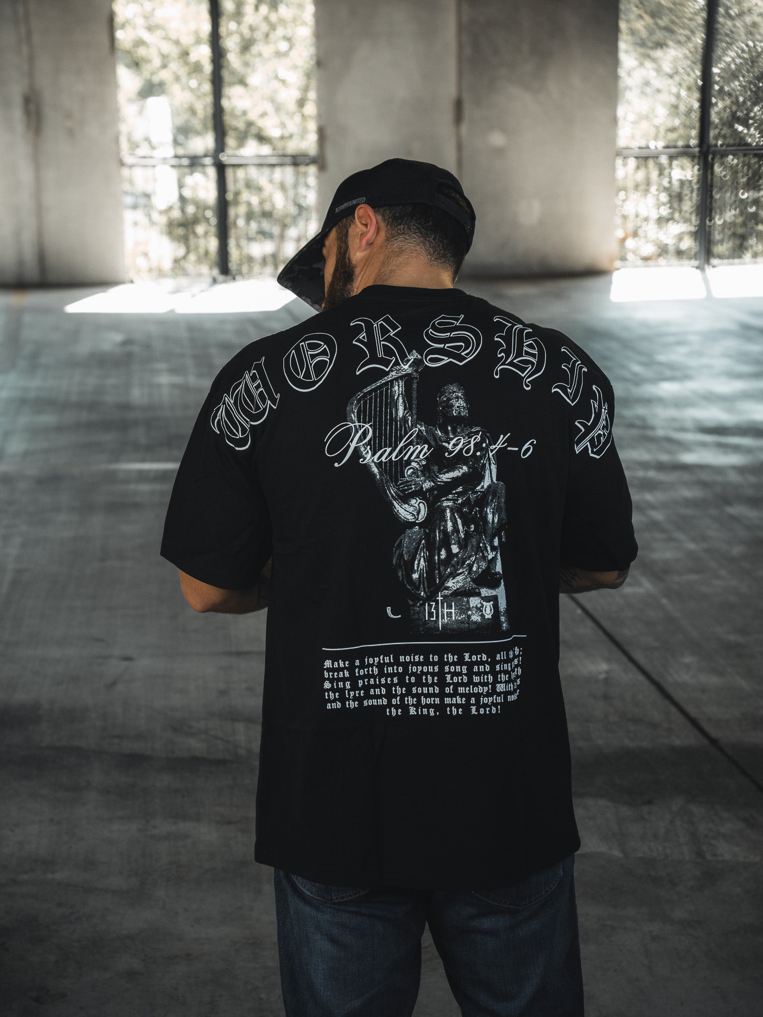 A man in a black cap and the 13th Unwavering Worship Premium Oversized Tee stands with his back to the camera. The shirt displays "WORSHIP," Psalm 98:4-6, and an illustration as sunlight streams through the windows ahead.