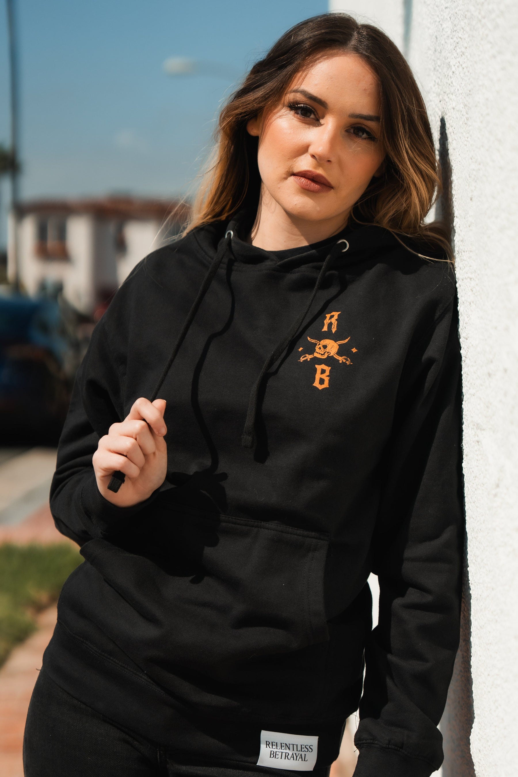 A woman with long hair in a black Standfast Gold Foil Hoodie by Relentless Betrayal leans against a white wall. Made from a cozy cotton/polyester blend, this unisex hoodie suits the sunny day as buildings and streets softly blur in the background.