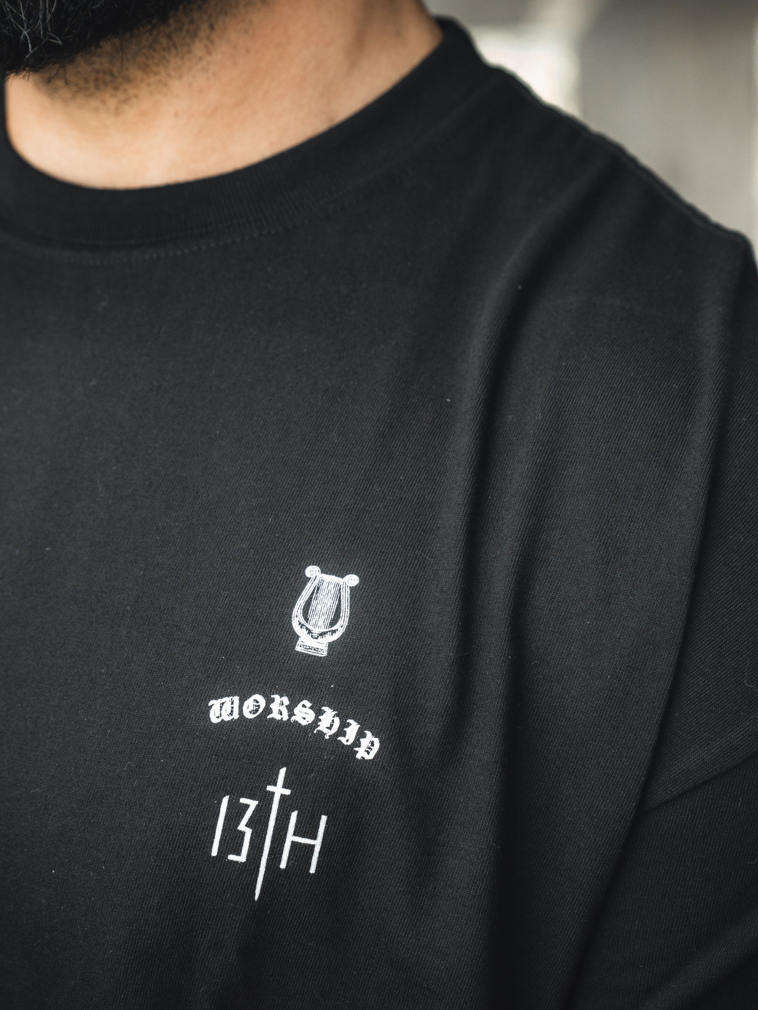 Close-up of a person in the Unwavering Worship - Premium Oversized Tee by 13th, featuring white designs—a small harp above "WORSHIP," “13TH,” and a cross-like symbol below. Inspired by Psalm 98:4-6. The person's face is not shown.