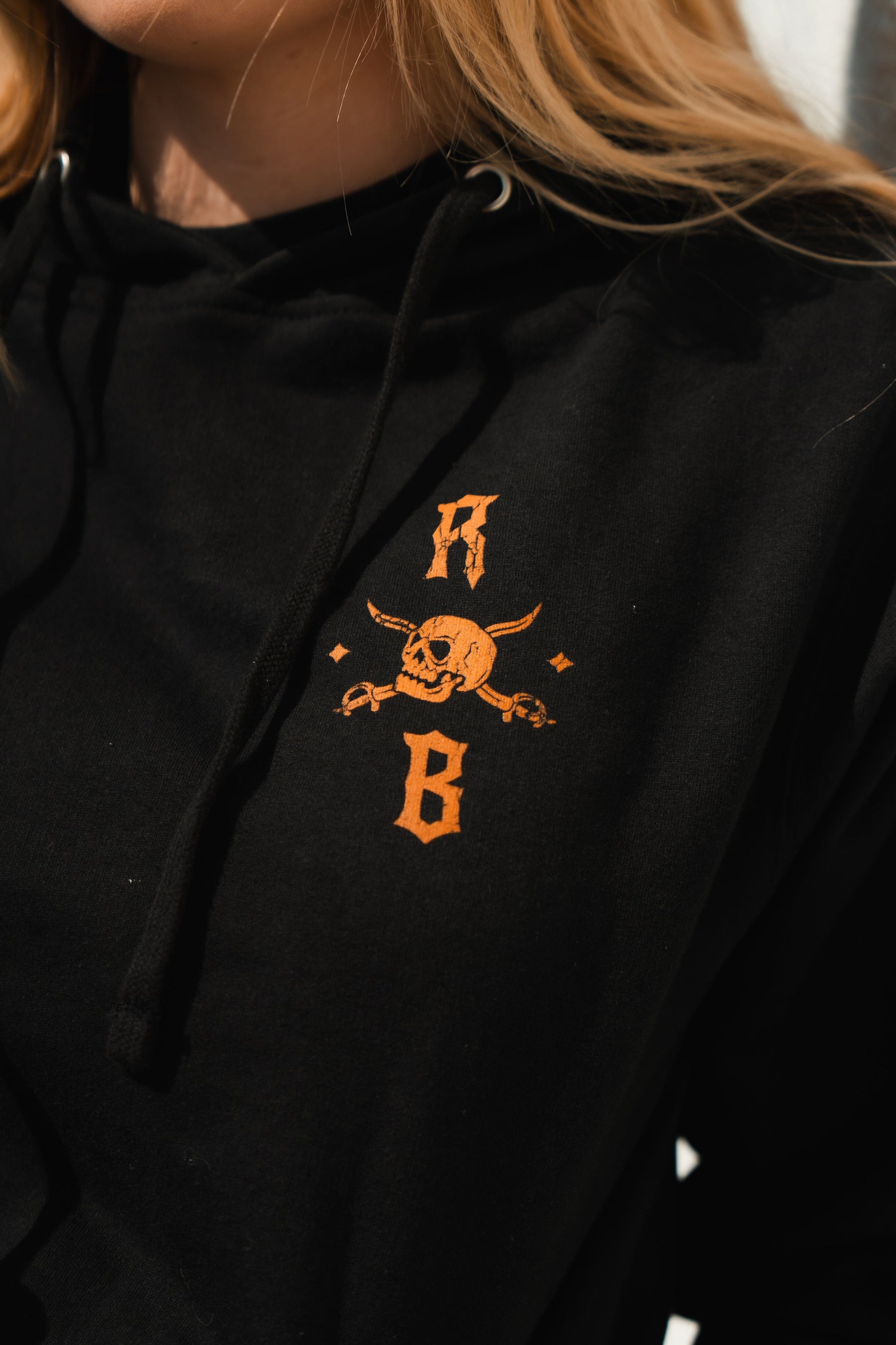 A close-up of someone wearing a Relentless Betrayal Standfast Gold Foil Hoodie features a bold orange skull and horns with crossed bones on the chest. The unisex black cotton/polyester hoodie subtly shows W and B amid their long hair.