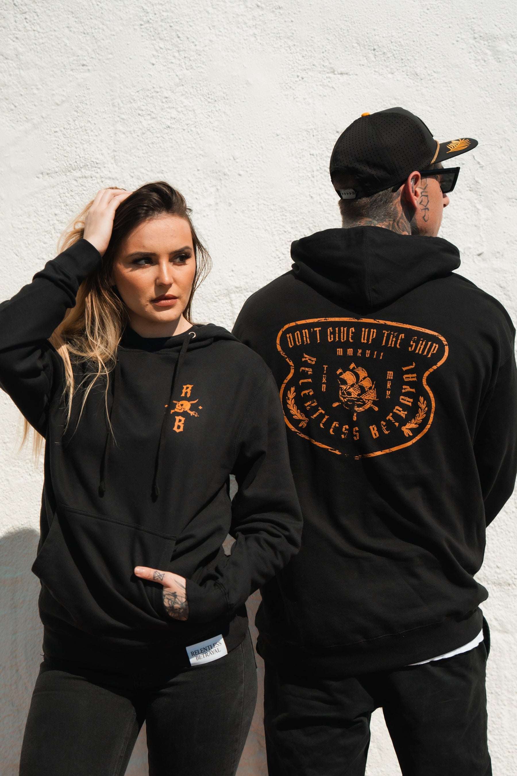 A woman and a man in black Standfast Gold Foil Hoodies by Relentless Betrayal stand against a white wall. The womans features an orange emblem, while the mans showcases Dont give up the ship. Both tattooed, they confidently wear these unisex cotton/polyester blend pieces.