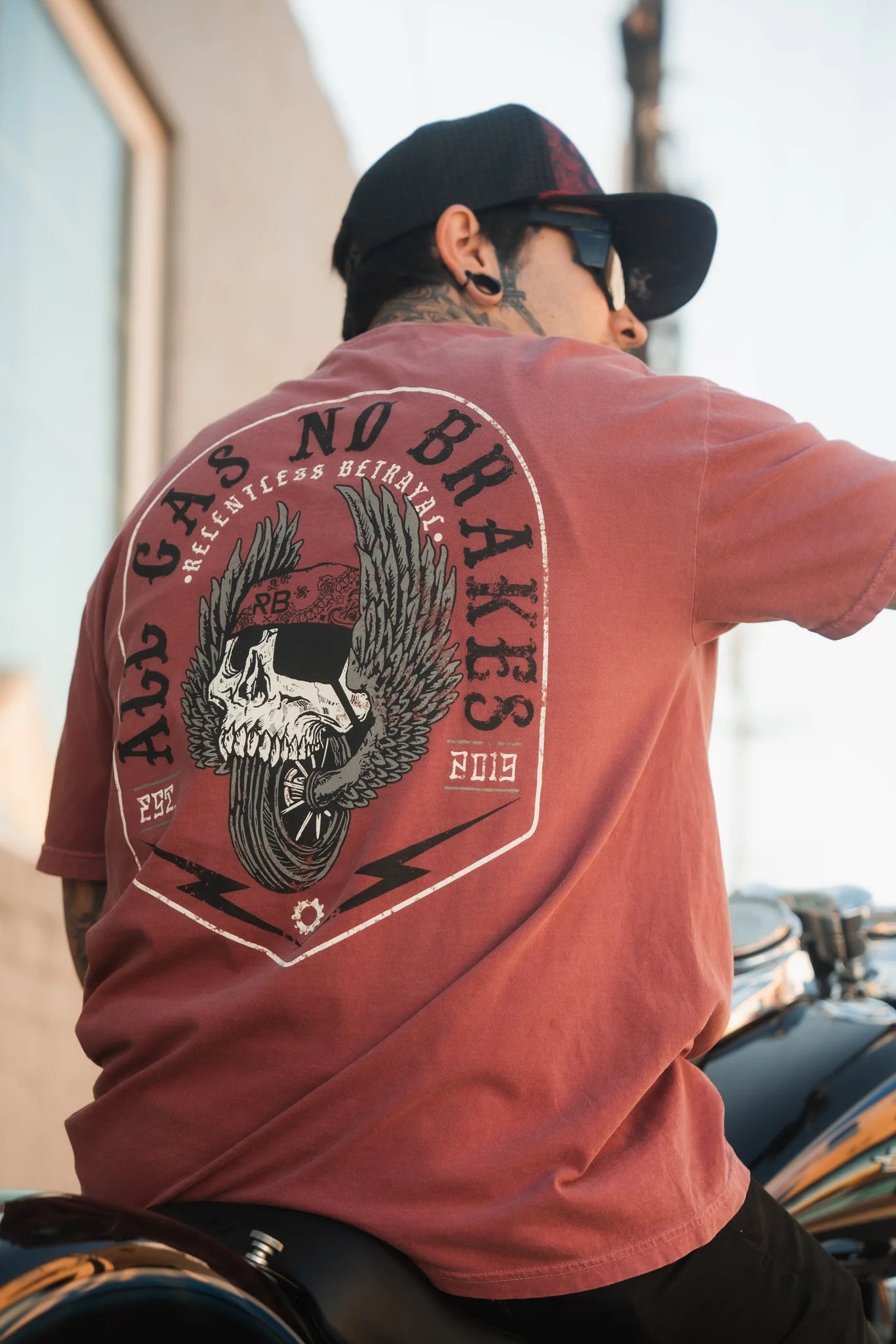 A man in a Relentless Betrayal All Gas No Brakes Premium Brick T-Shirt with a skull, wings, and No Brakes design sits on a motorcycle exuding youthful fearlessness. Hes wearing sunglasses and a cap as he gazes into the distance.
