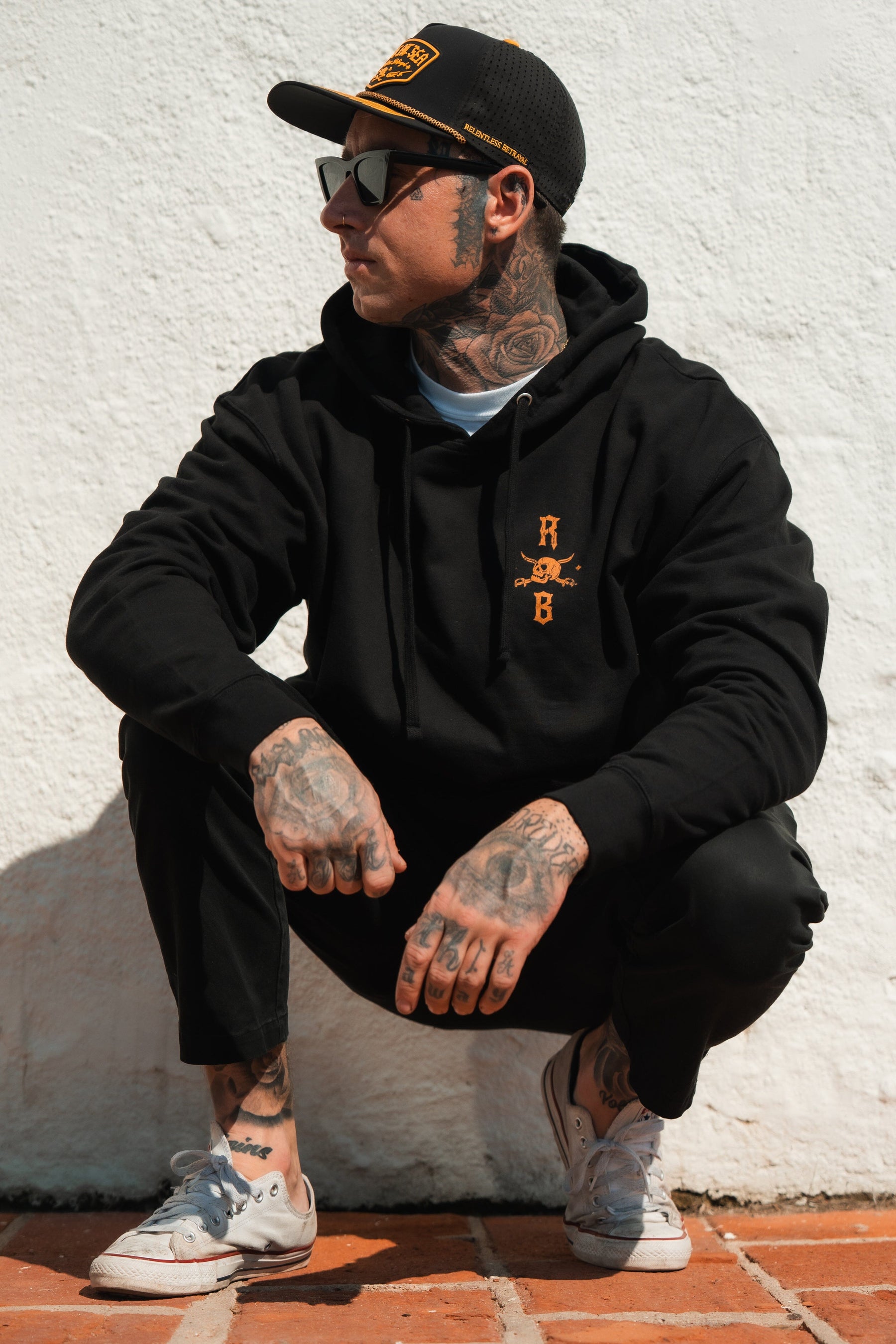 A person wears a black, unisex Standfast Gold Foil Hoodie by Relentless Betrayal made from a cotton/polyester blend while crouching against a white wall. They show off tattoos and finish their casual style with a cap, sunglasses, and white sneakers.