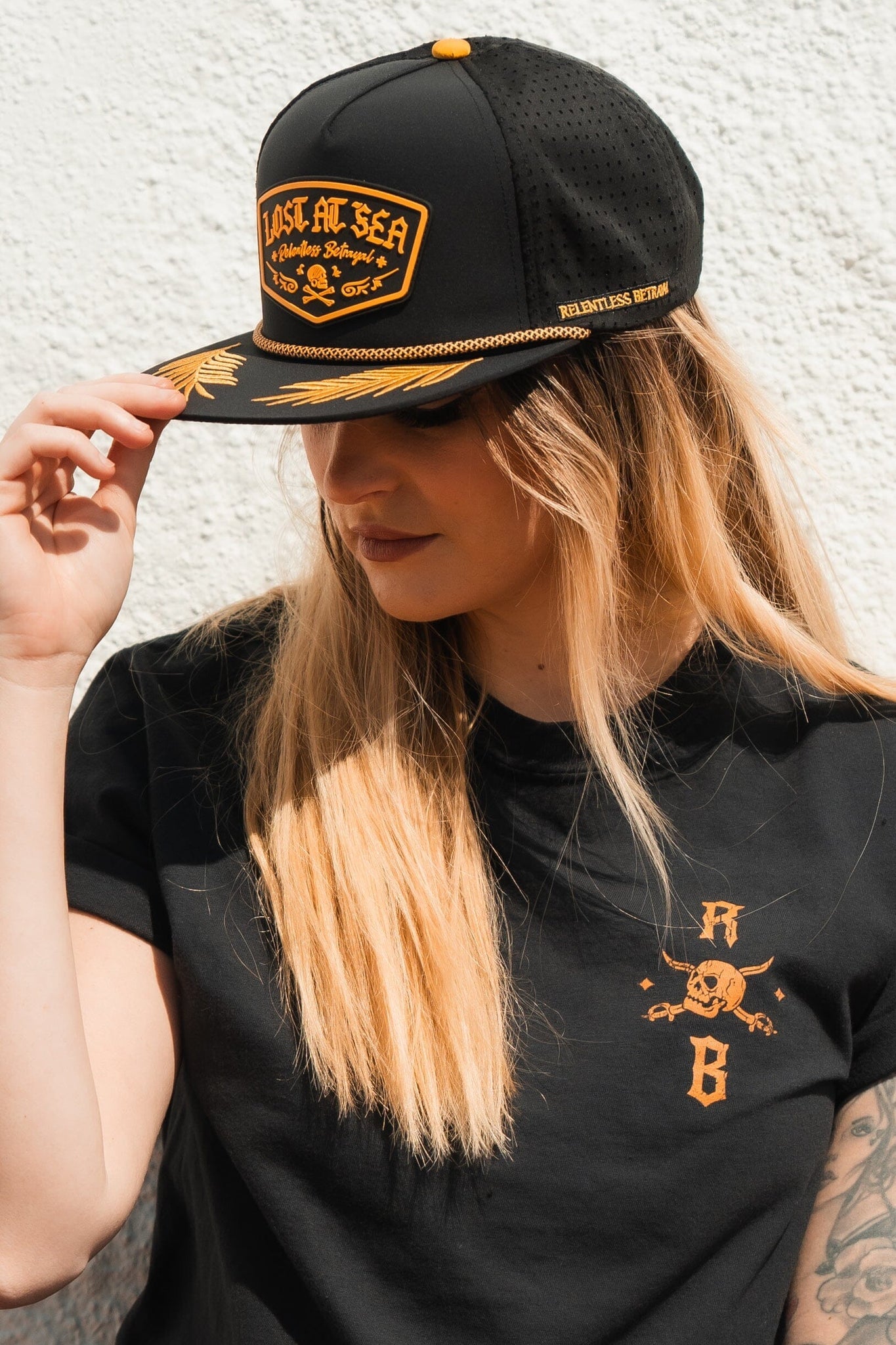 A person with long hair dons the Relentless Betrayals Lost At Sea PVC Snapback in black and gold, embodying the pirate lifestyle. They wear a black shirt with gold embroidery, adjusting their water-resistant cap against a light background.
