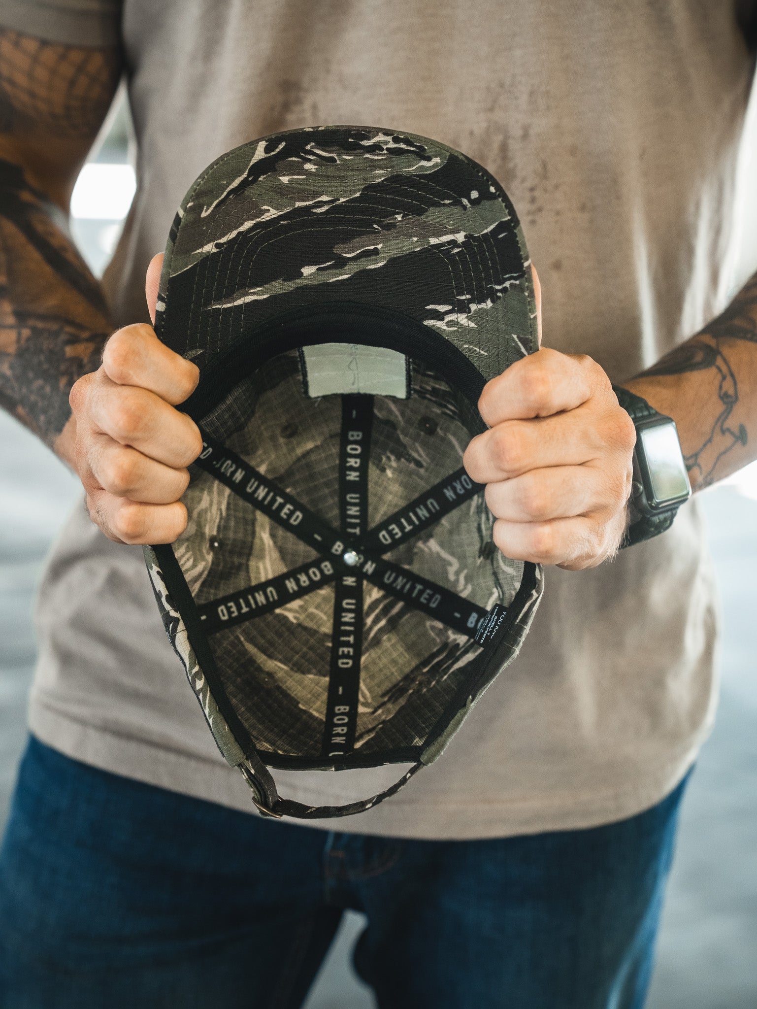 A tattooed individual holds the Born United Buckle Hat - Savage Operations by Born United Private Label, displaying its interior black straps labeled BORN UNITED. They wear a gray shirt, blue jeans, and a black wristwatch.
