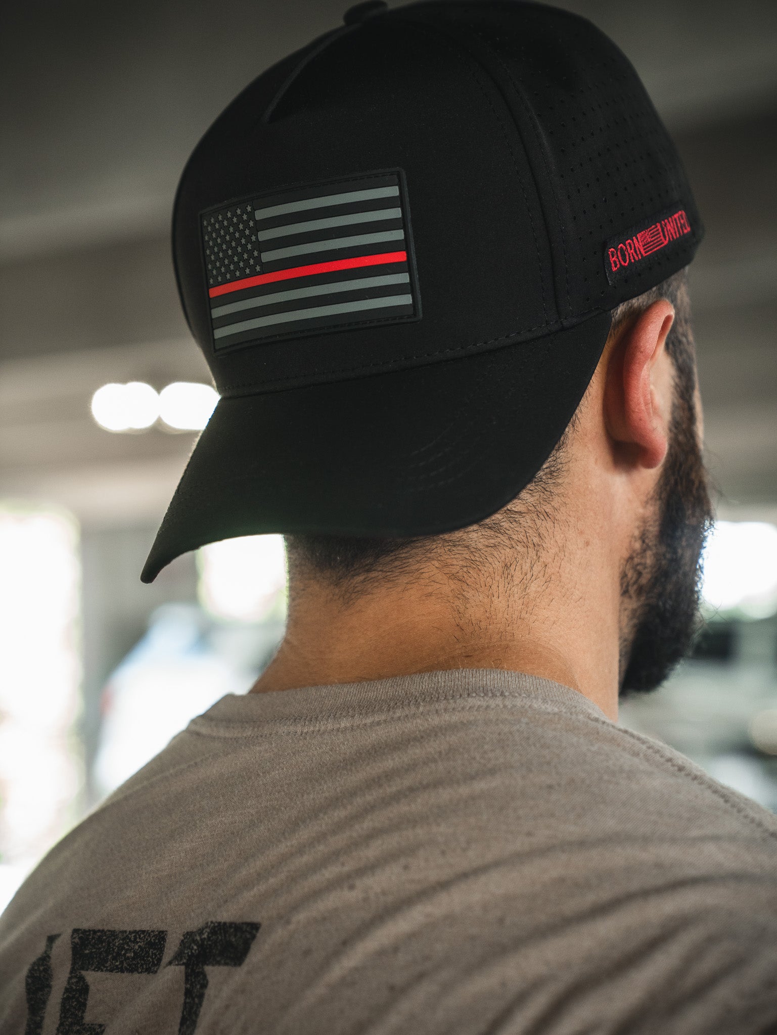 A man in a light gray shirt is seen from behind wearing the Born United Premium Flag Snapback - TRL by Born United Private Label, featuring a black cap with a subdued flag patch, red stripe, and "BORN PRIMITIVE" on the side.